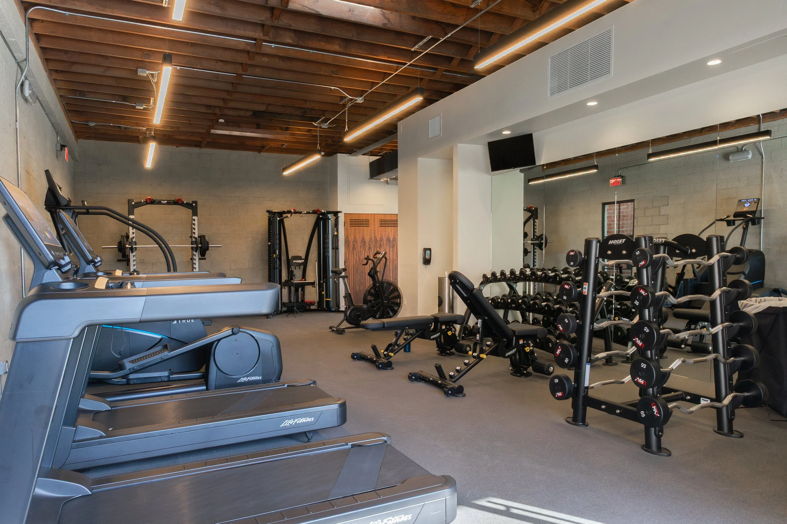 Modern gym with cardio machines, weight racks, and exercise equipment in a well-lit space with wooden ceiling beams and concrete walls.