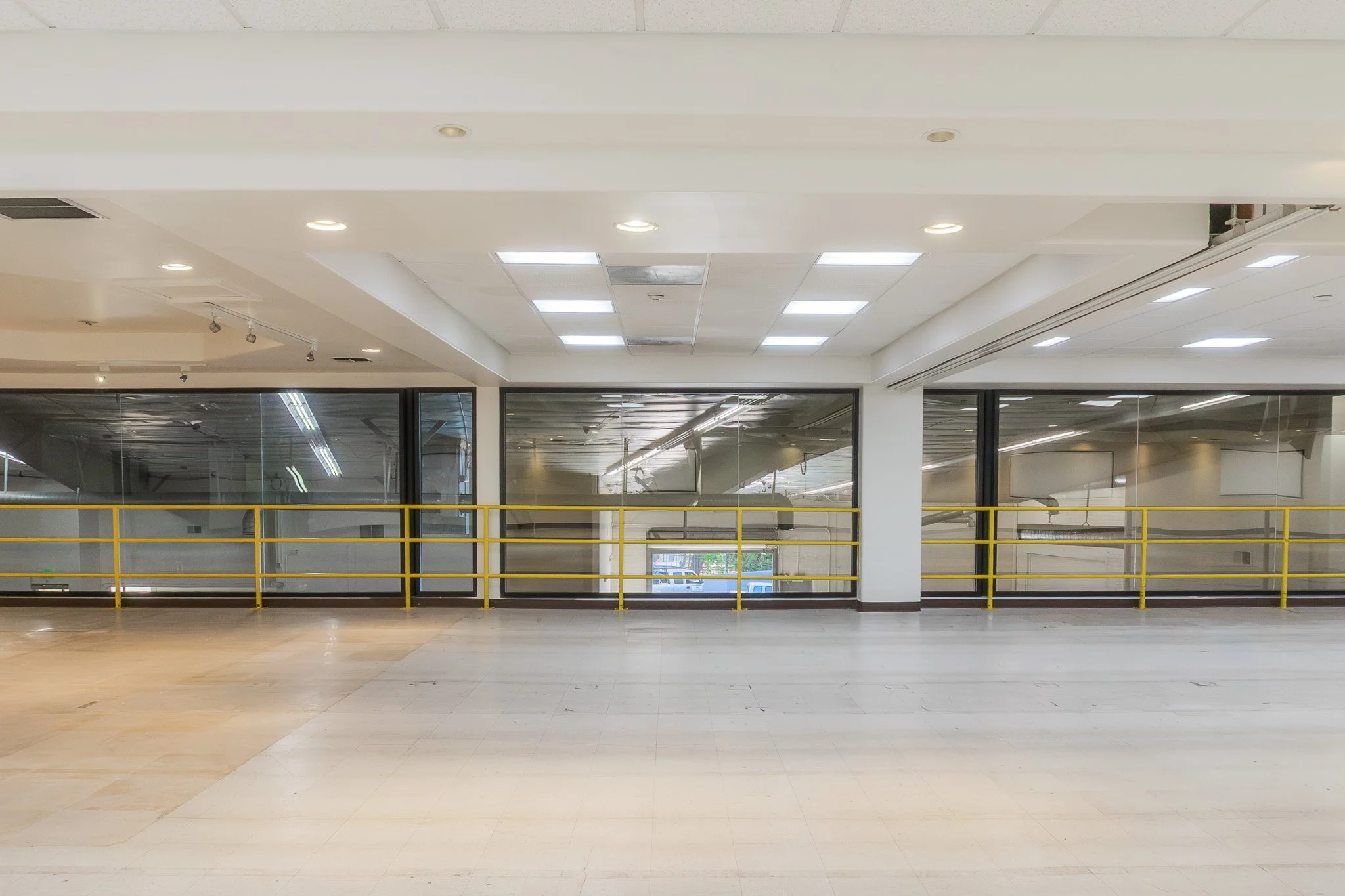 Empty indoor space with large glass windows, white ceiling with lights, beige floor, and yellow safety rail in the background.