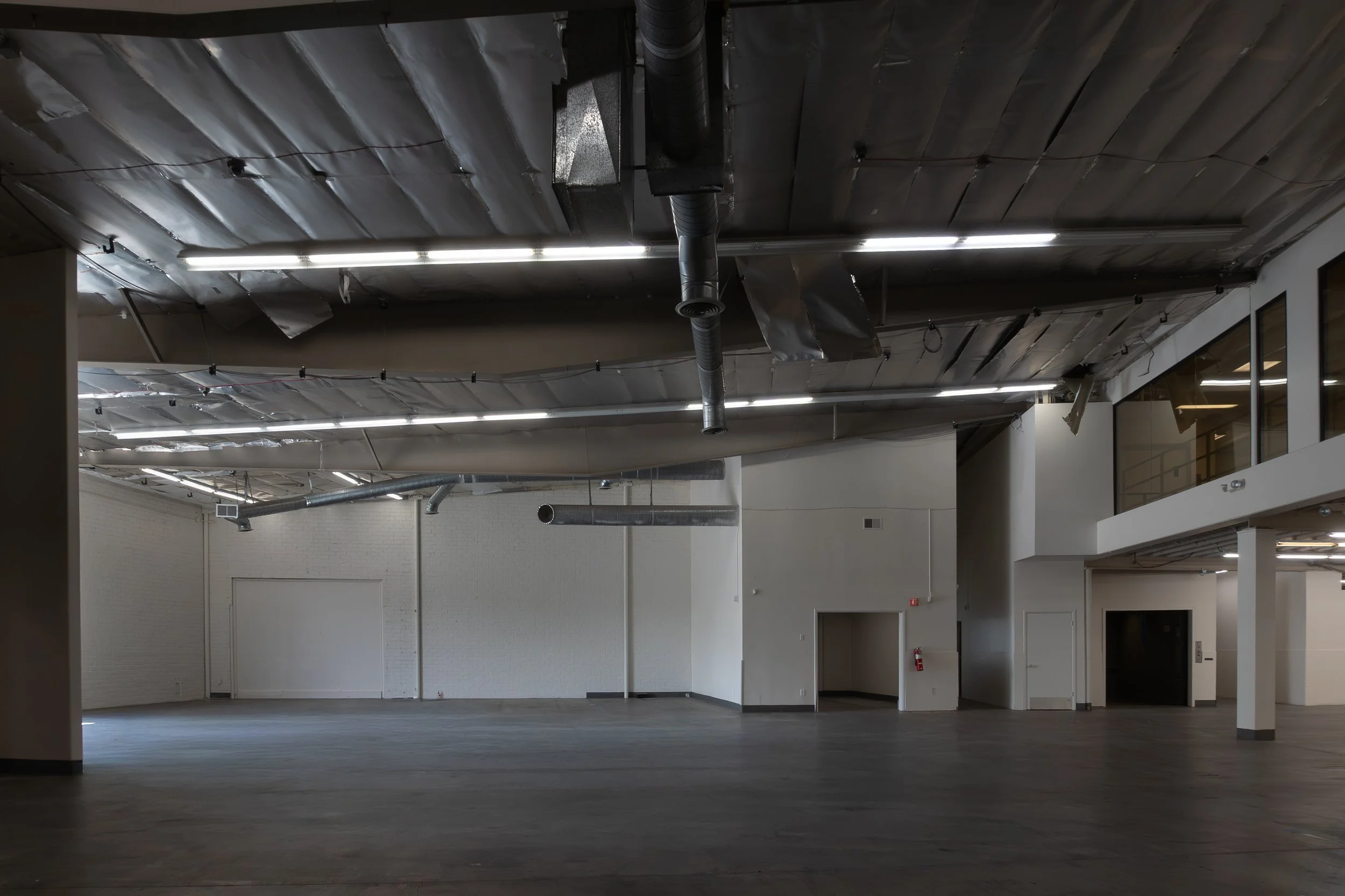Empty commercial or industrial space with unfinished ceiling showing exposed pipes and ductwork, white walls, and wood-look flooring.