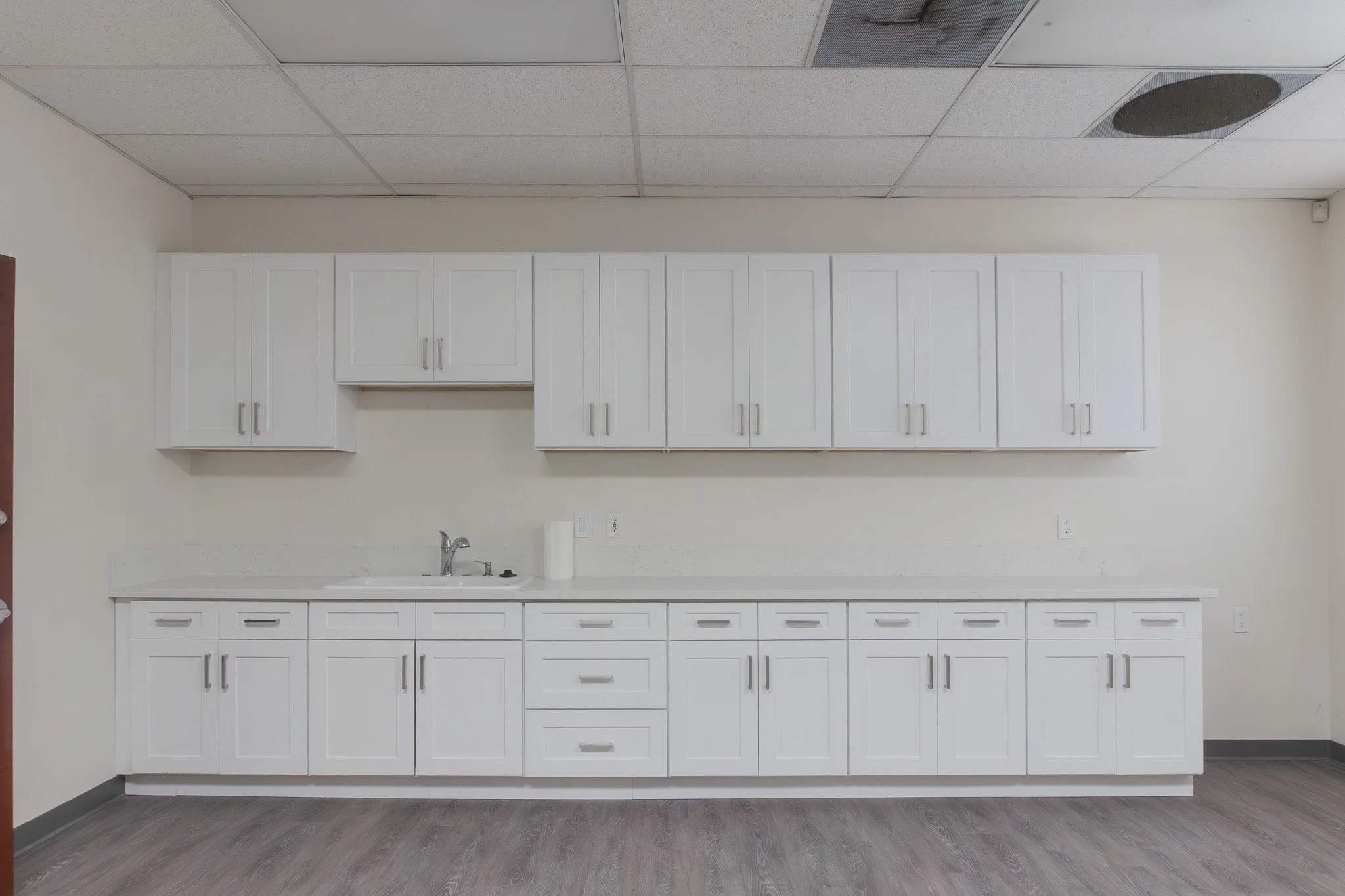 Empty white kitchen cabinets with countertop, sink, and faucet in a room with ceiling tiles and wood-like flooring.