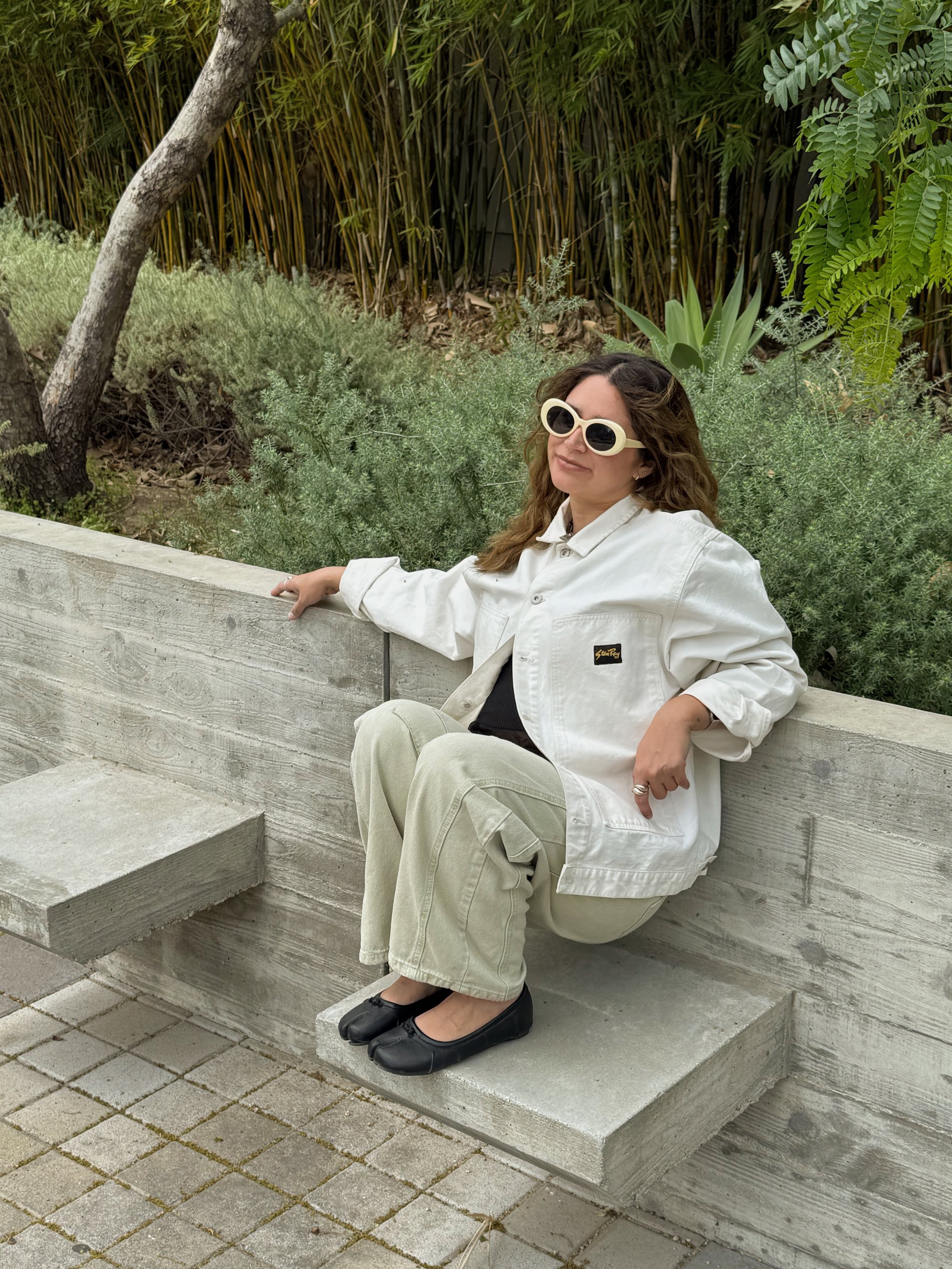 Woman sitting on a concrete bench outdoors, wearing white jacket, beige pants, black shoes, and large white sunglasses, with greenery in the background.