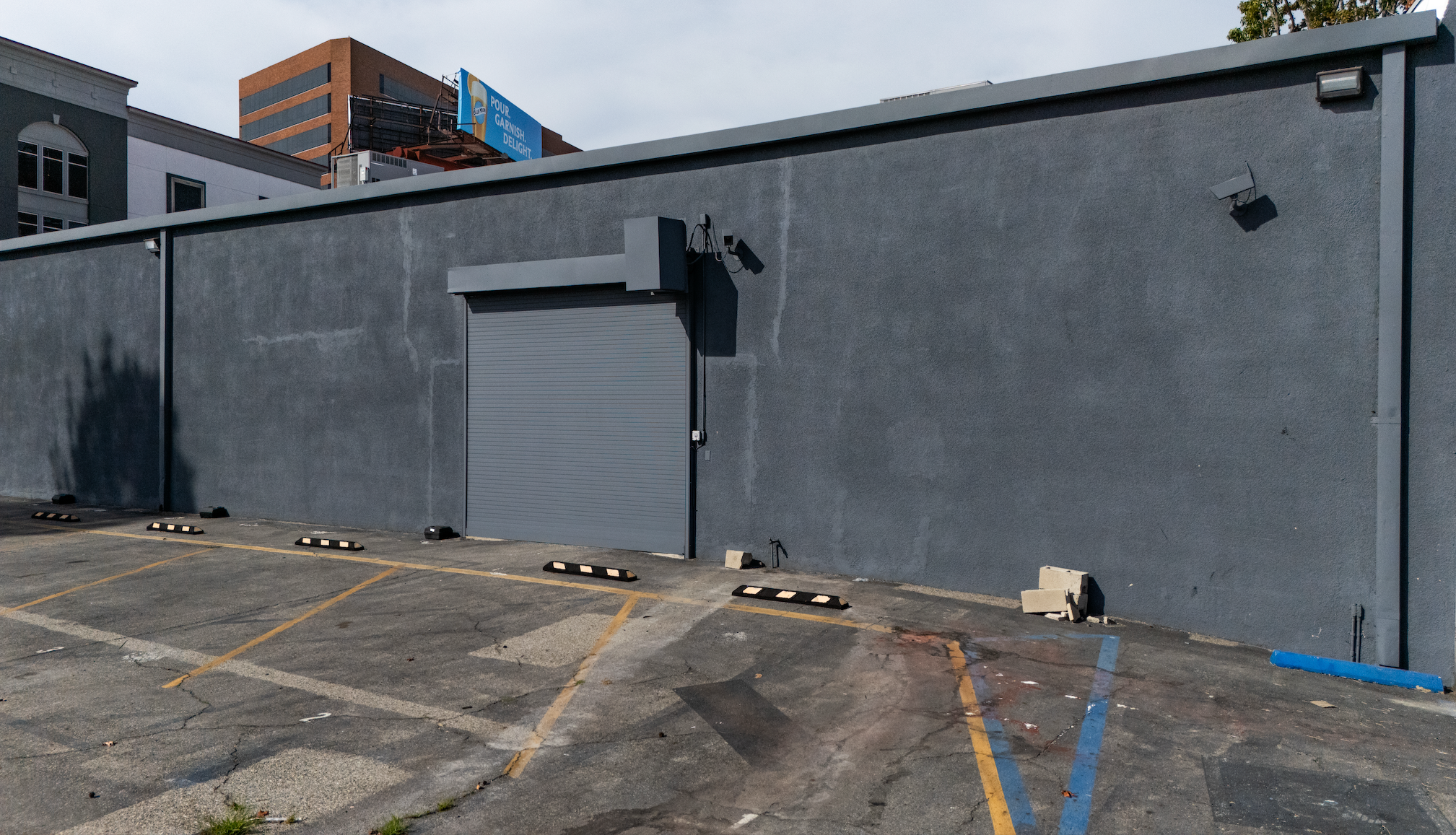 An empty parking lot with yellow lines, wheel stops, and a gray wall with a closed gray roll-up door, two security cameras, a small pile of bricks, and a blue pipe, with buildings visible in the background under an overcast sky.