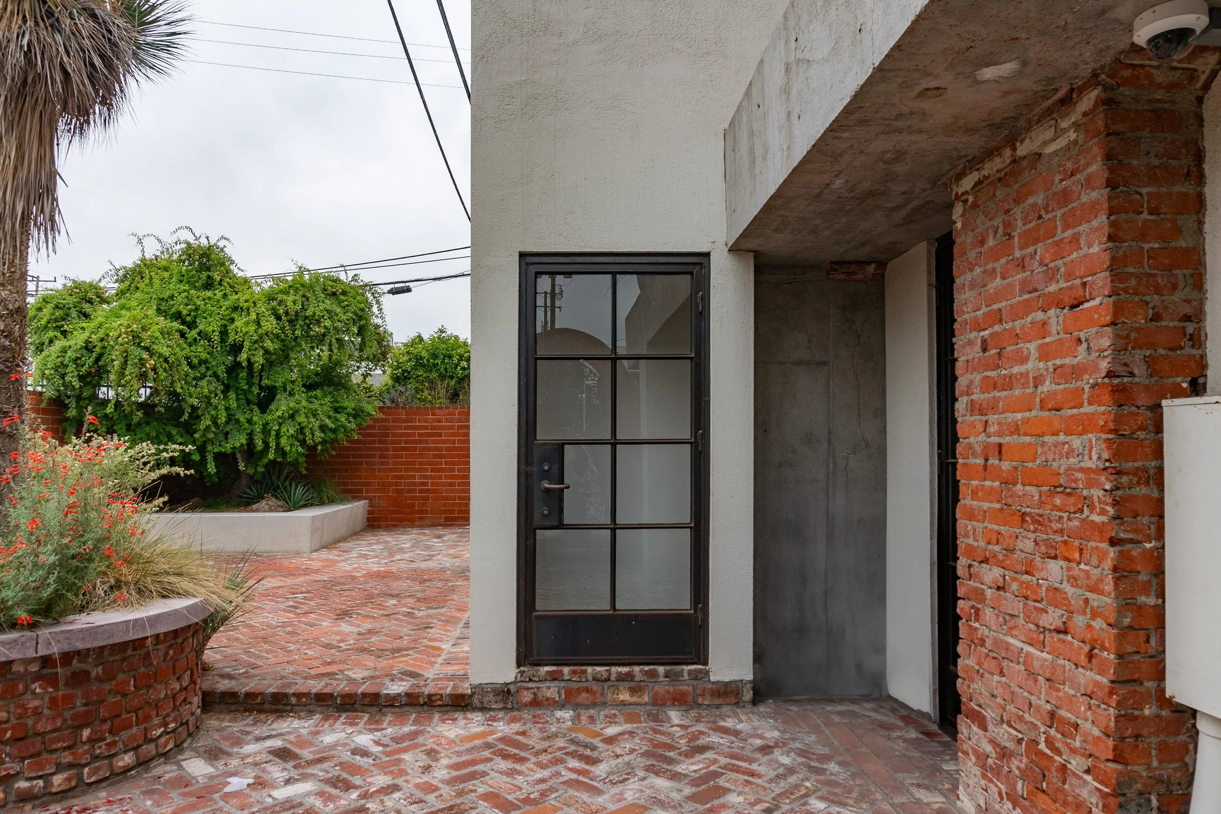 Exterior of a building with a black metal door and brick wall, brick pavement, and a garden with green bushes and plants.