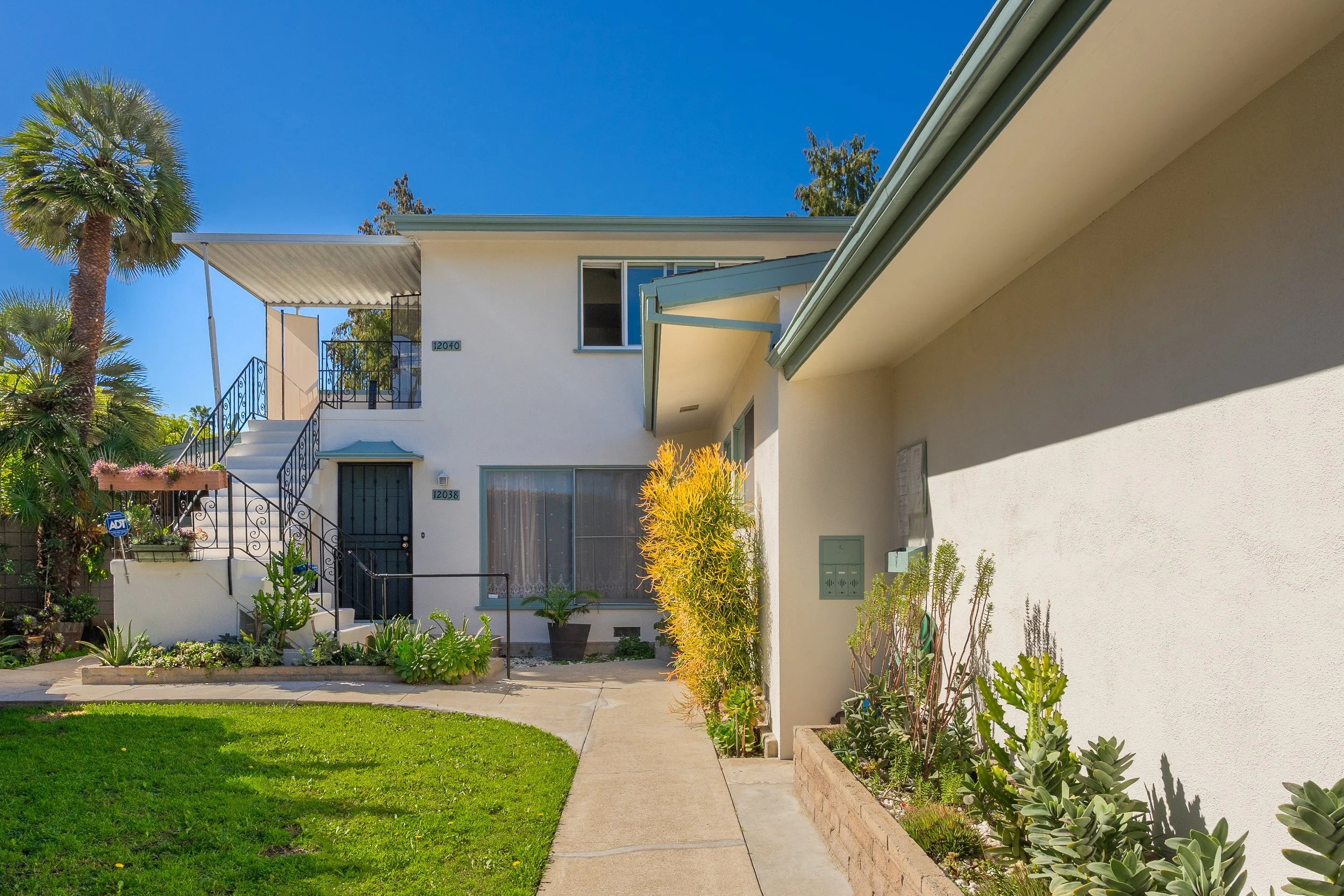 Exterior view of a modern two-story residential building with white walls, black metal staircase, and stairs leading to the upper floor, surrounded by green plants, palm trees, and a bright blue sky.