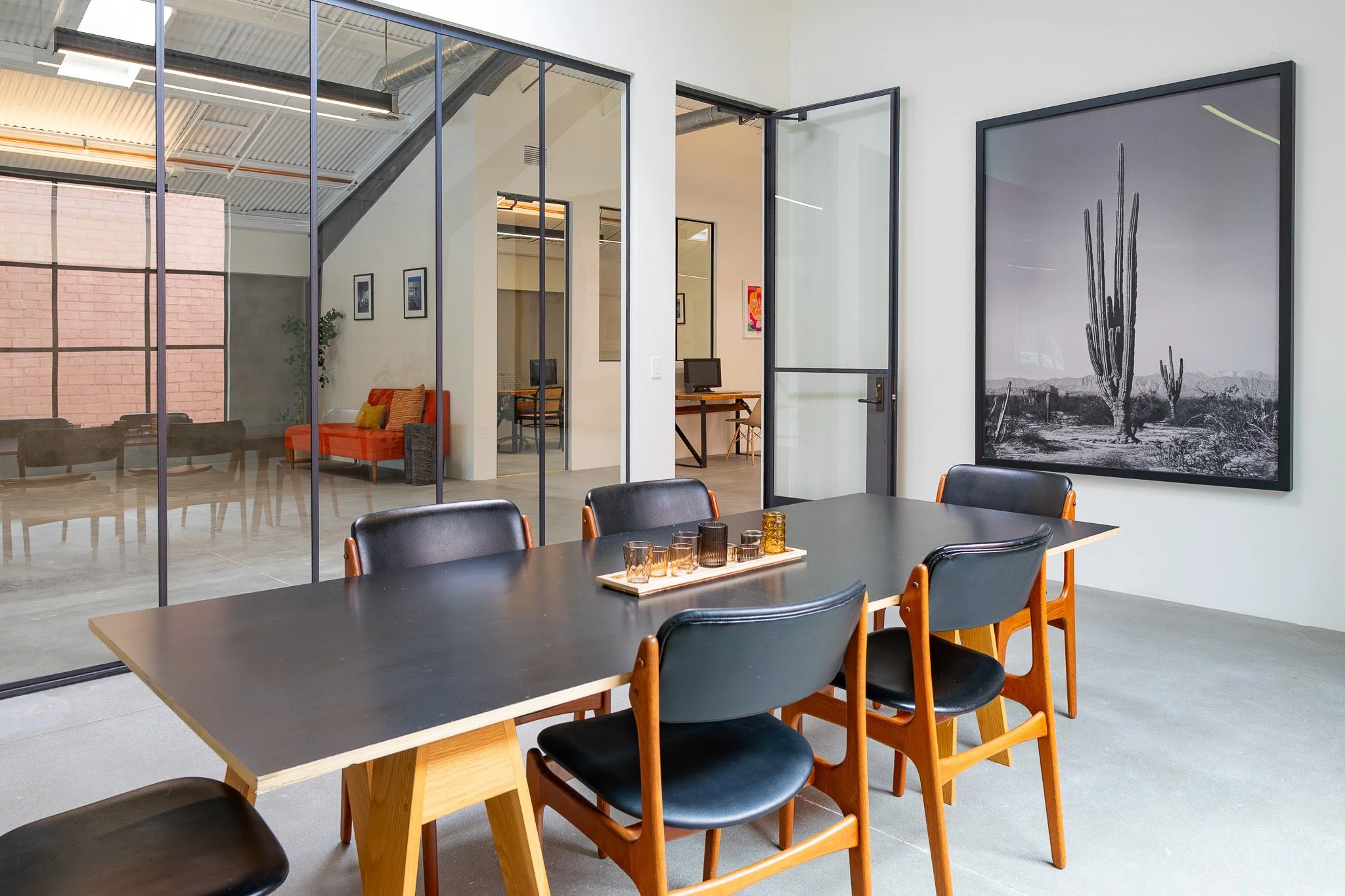 Modern office or conference room with a large black table and black chairs, decor including a large black and white cactus print on the wall, and glass walls separating different work areas.