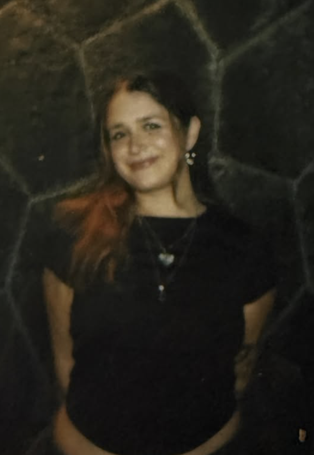 A woman with dark hair and earrings smiling, standing in front of a stone wall at night.