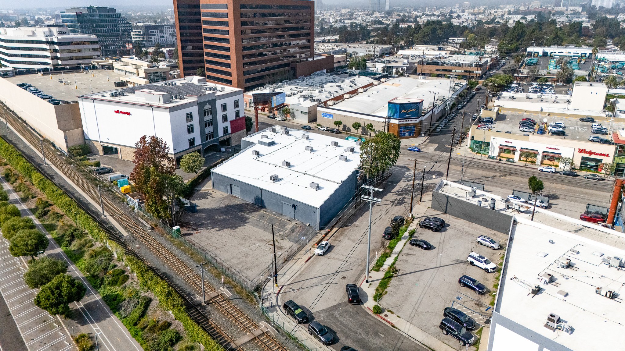 Aerial view of an urban area showing buildings, parking lots, and a railway track with trees along the side.