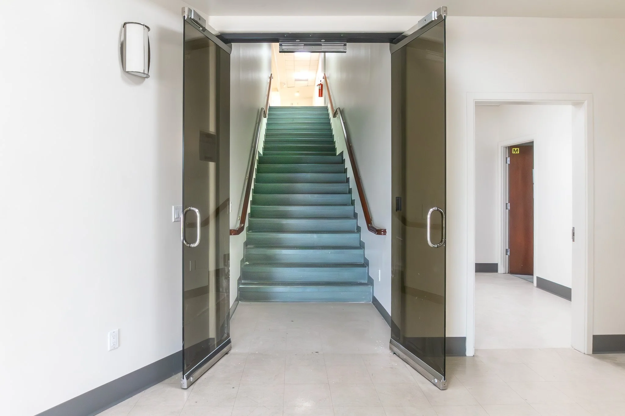 Glass doors open to a hallway with a stairway leading upward, with green stairs and wooden handrails. There is a wall-mounted device on the left wall and a door to the right of the stairway.