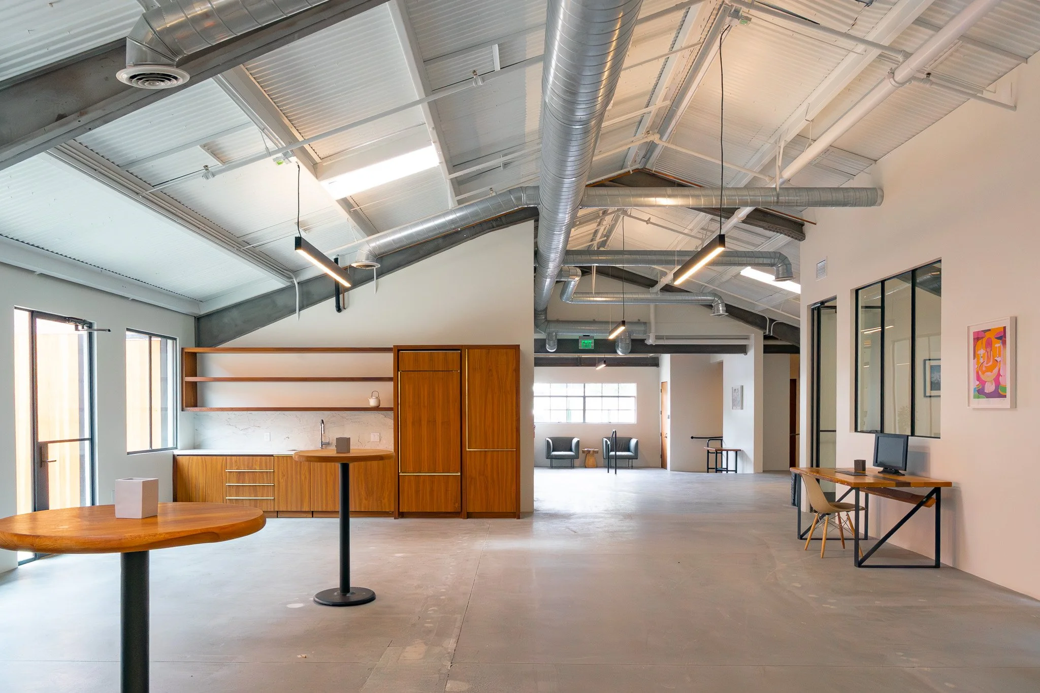 Open-plan office with modern industrial ceiling, large windows, wooden furniture, and a seating area.