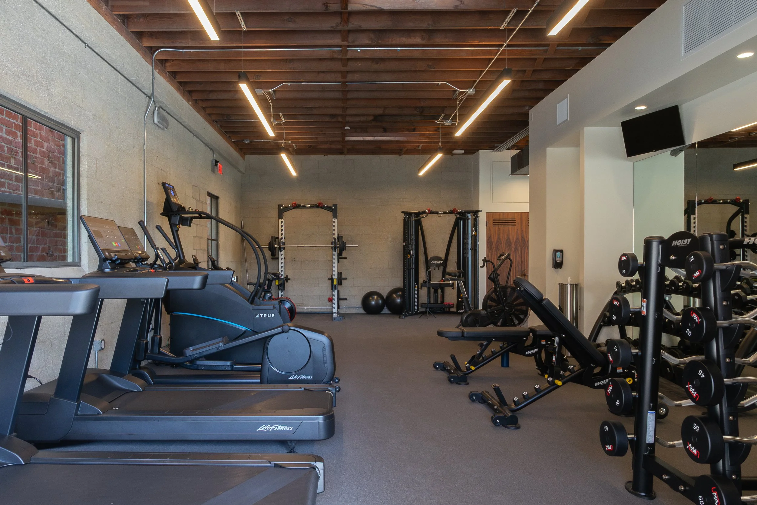 Interior view of a modern gym with various exercise equipment including treadmills, ellipticals, weight machines, dumbbells, exercise balls, and benches, with a wooden ceiling and brick and cinder block walls.