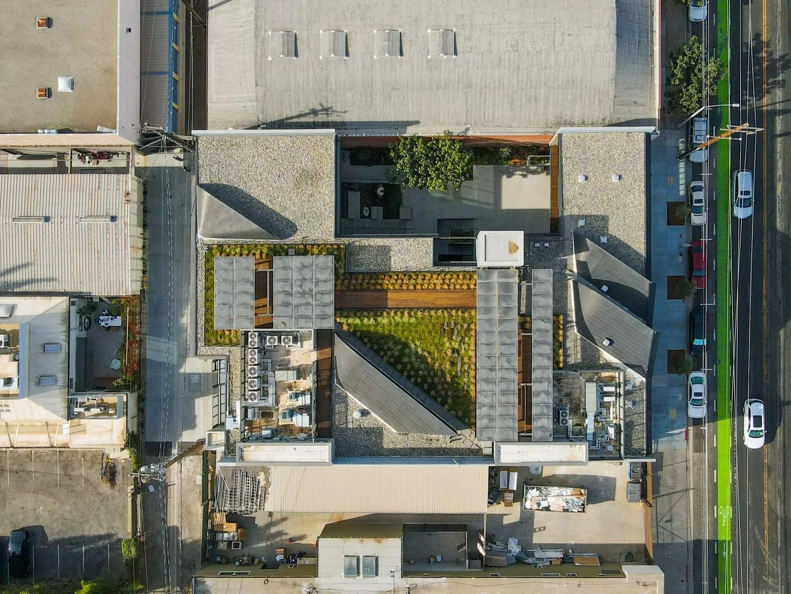 An aerial view of a city building rooftop with a garden, HVAC units, and adjacent parking lot and street.
