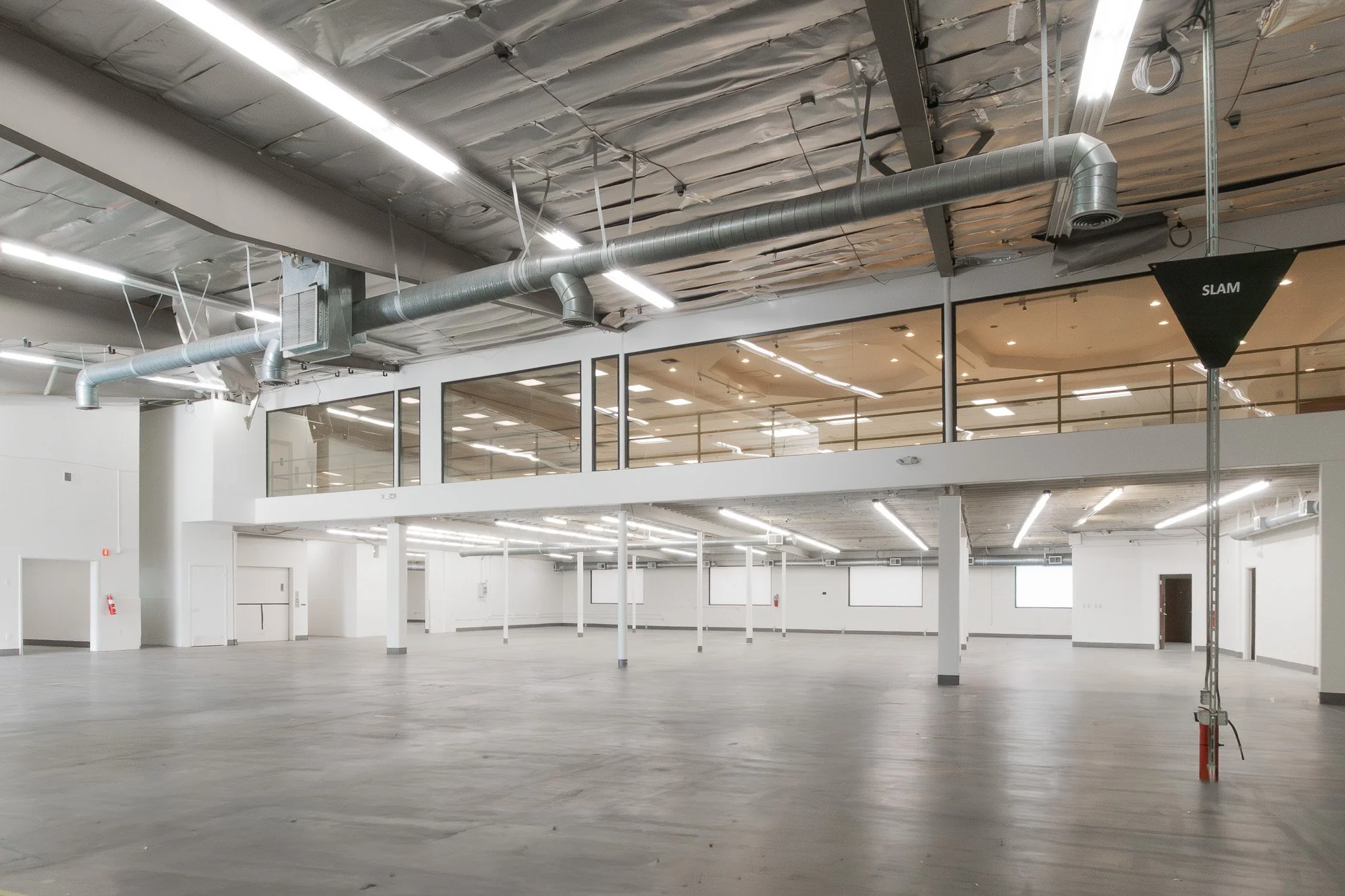 Empty, industrial-style commercial or office space with exposed ceiling ducts, large windows, and a mezzanine level.