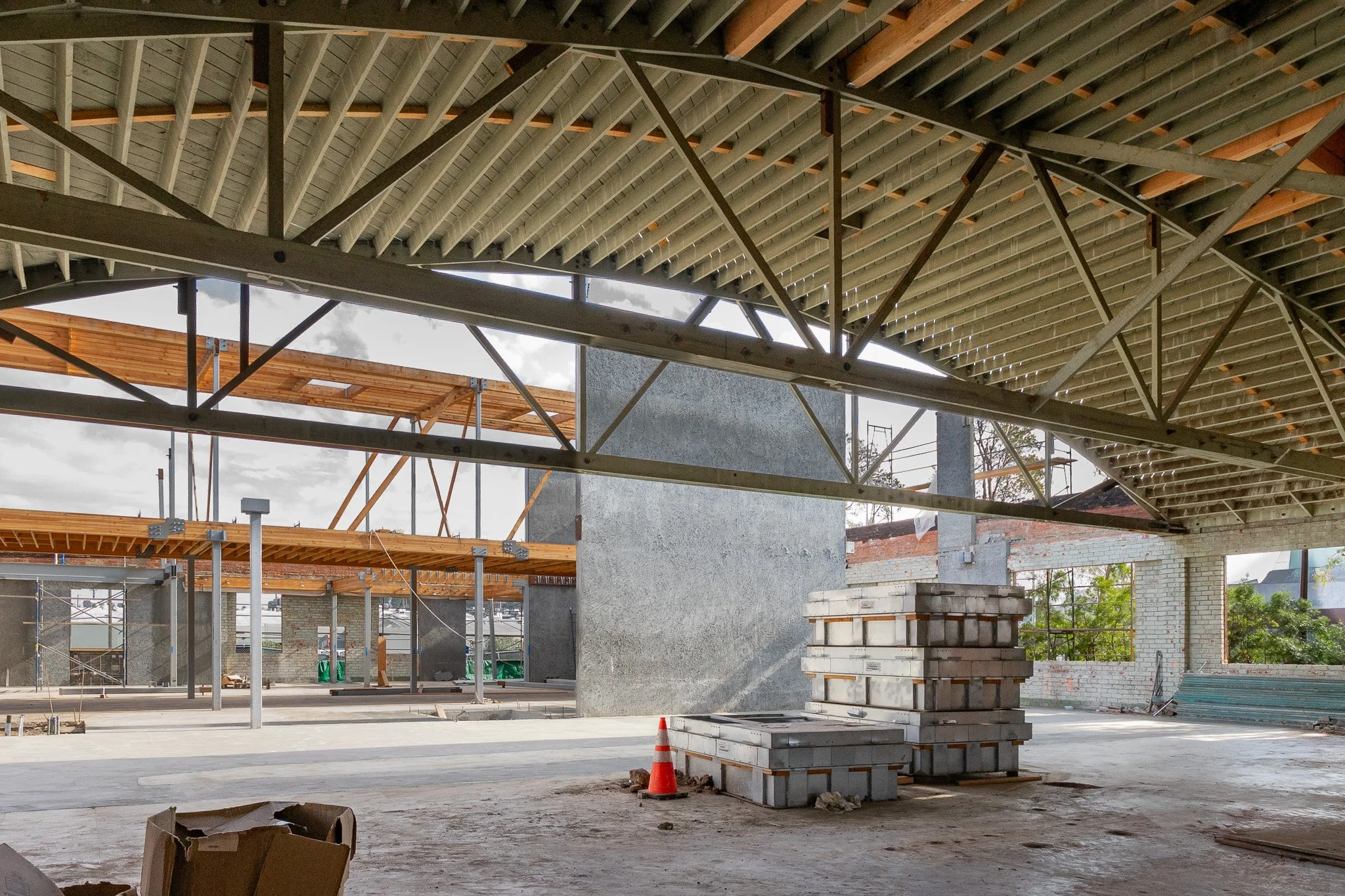 Construction site indoors with steel beams, stacks of bricks, and orange safety cones. Partially built walls and scaffolding are visible.