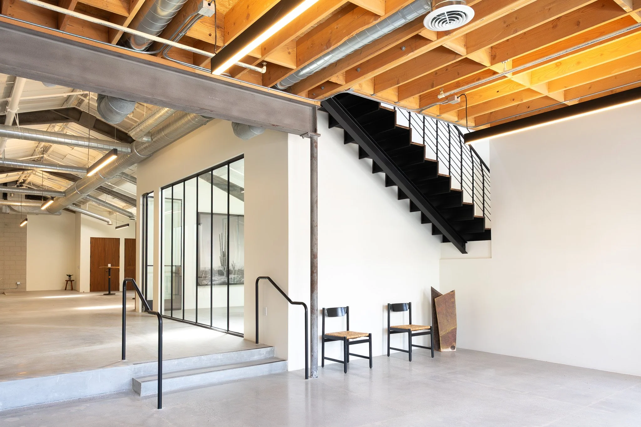 Interior of a modern building with a staircase, exposed wooden ceiling beams, metallic ductwork, and large glass panels.