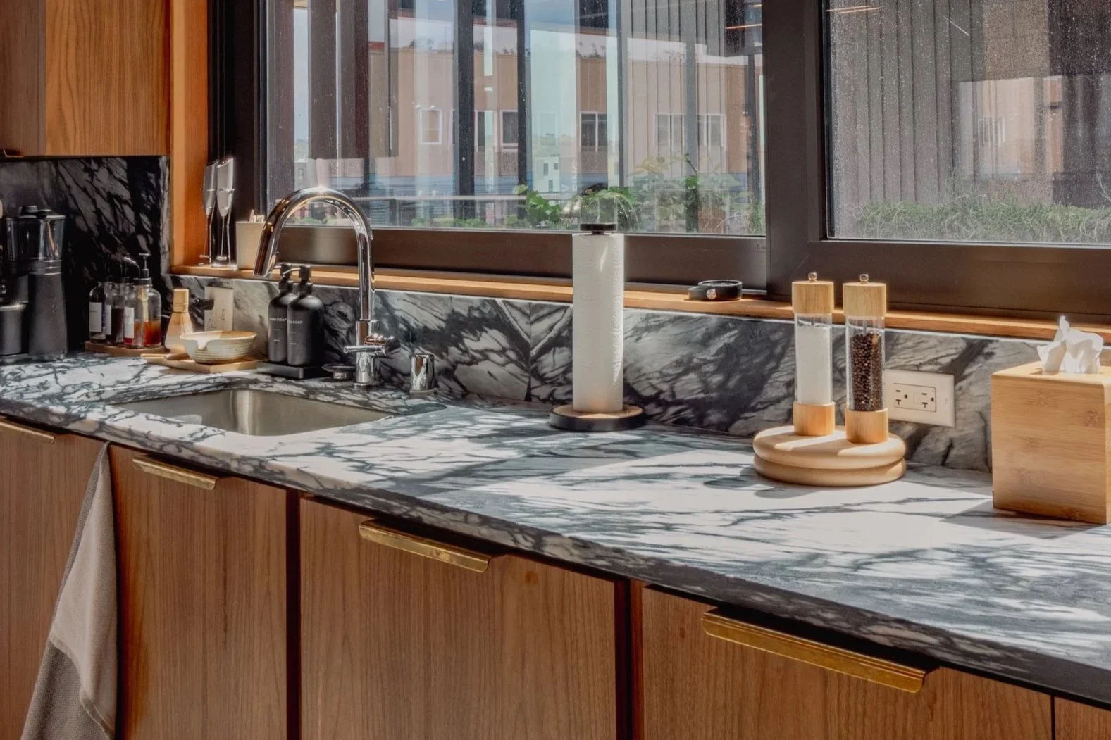 Kitchen countertop with black and white marble pattern, a window with a view outside, a sink with a faucet, a paper towel holder, salt and pepper shakers, and a tissue box.