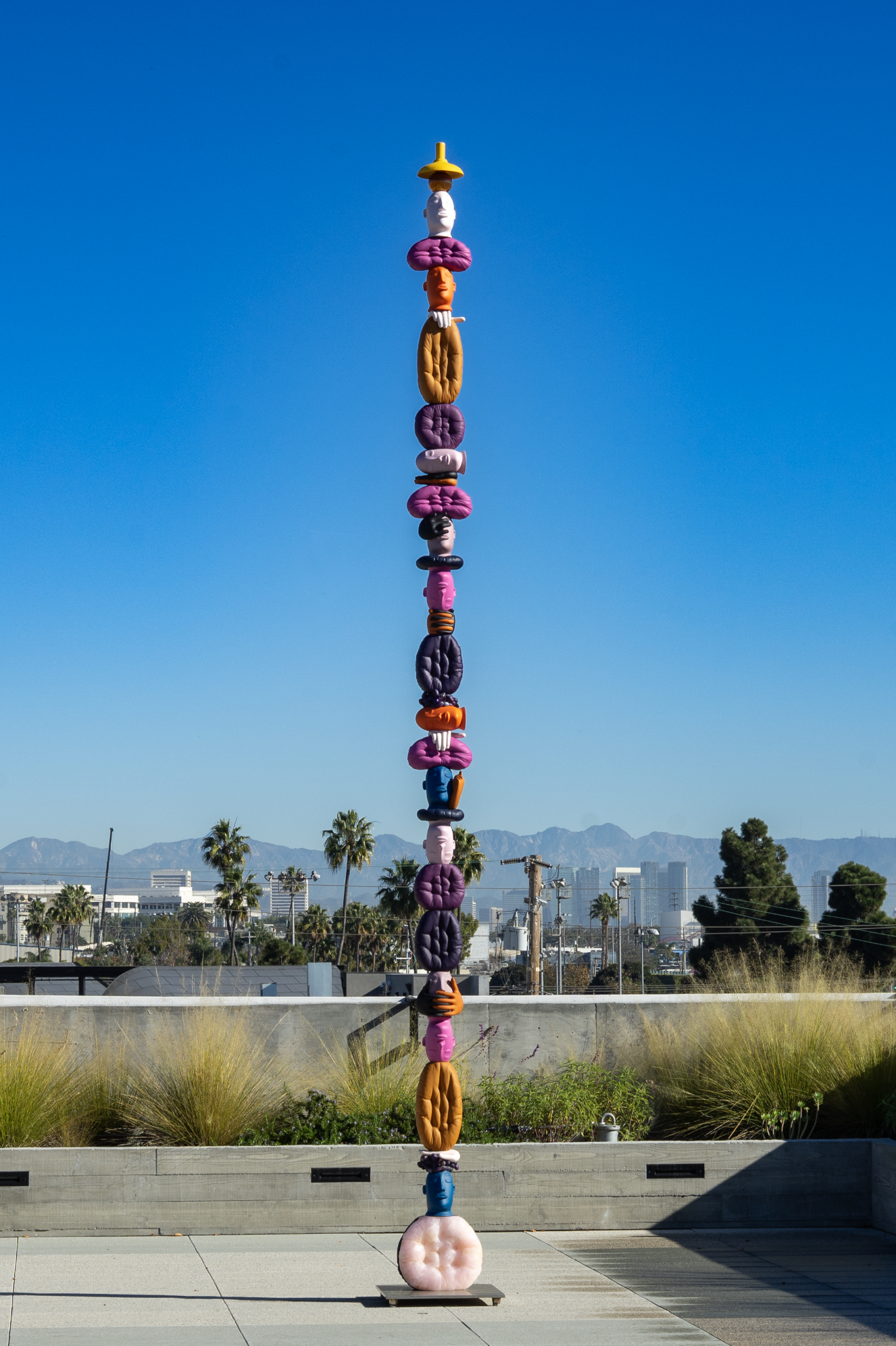 A tall outdoor sculpture made of stacked colorful bean bag chairs and objects against a clear blue sky with distant mountains and palm trees in the background.