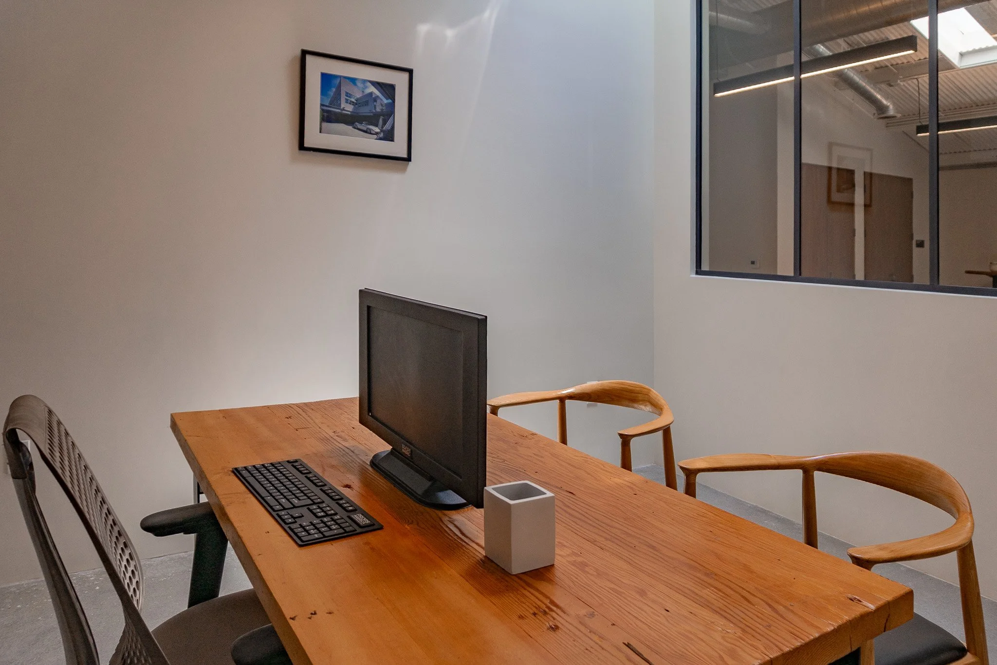 An office conference room with a wooden table, a computer monitor, a keyboard, a white square container, and three chairs. One chair is made of mesh and metal, and two are wooden with armrests. There is a framed picture of a building on the wall and 