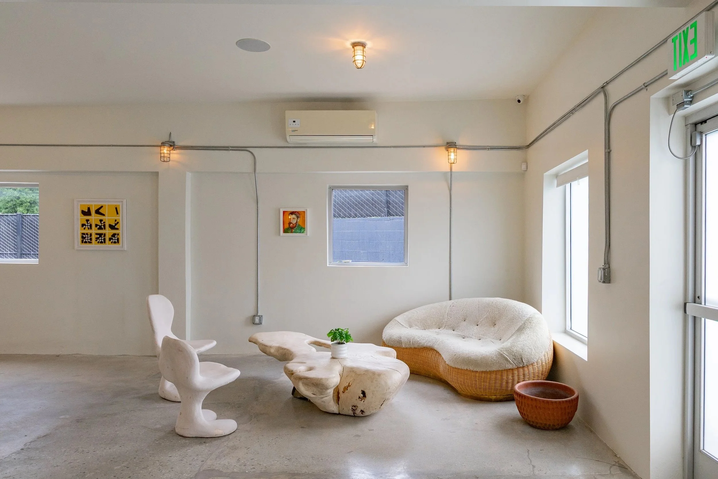 A minimalist living room with white walls, modern furniture, and abstract artwork, featuring a large curved sofa, unique wooden tables, and potted plants near windows, with industrial-style lighting and an exit sign.