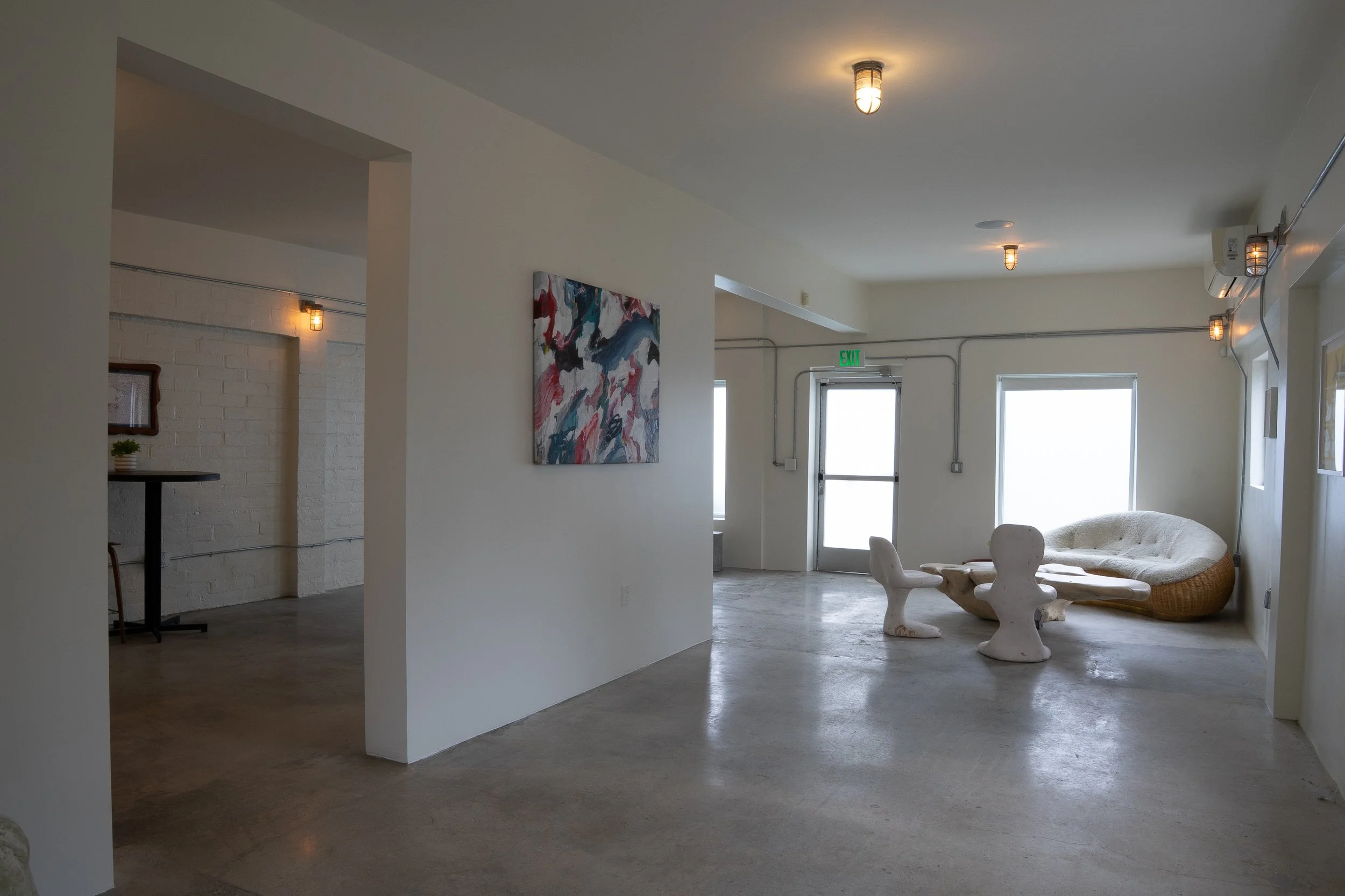 A minimalistic modern interior with white walls, concrete floors, abstract painting, and unique furniture including a textured sofa and sculptural chairs, near an exit door and large windows.