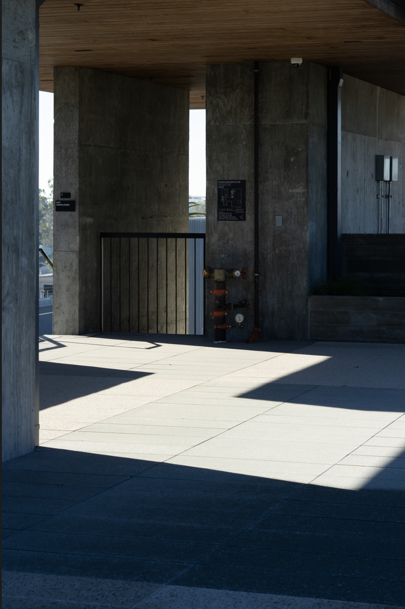 Concrete building exterior with a shaded area, a black railing, a fire pipe, and a framed evacuation plan, with sunlight casting shadows on the tiled ground.
