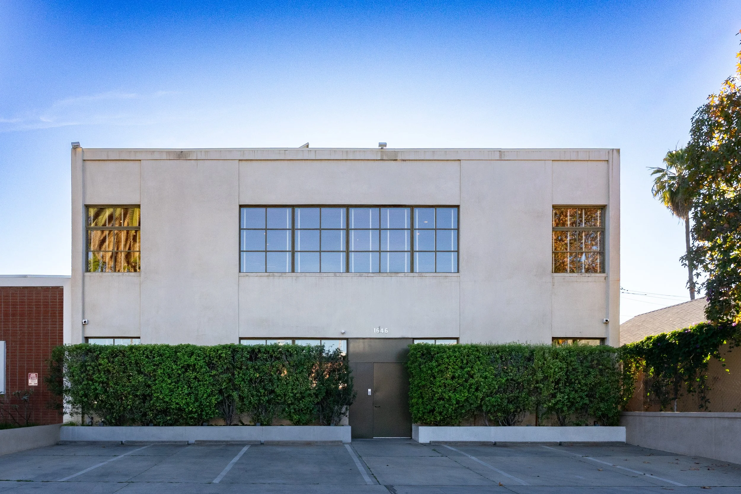 Modern two-story building with white exterior, large front windows, and green bushes in front, set against a clear blue sky.