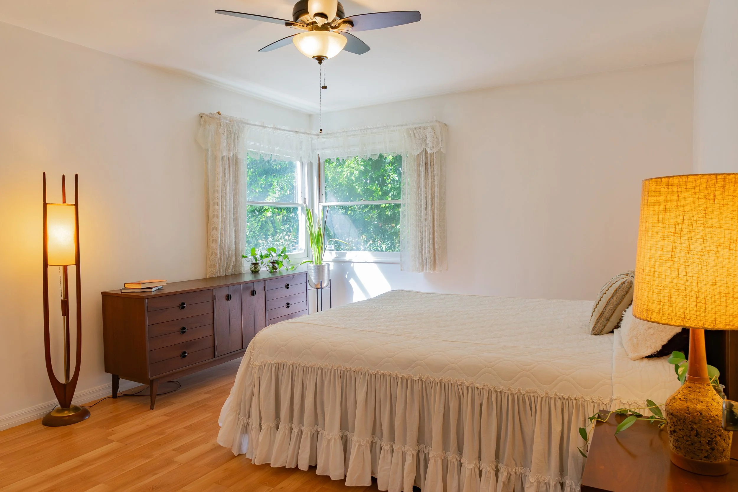 A bedroom with a wooden dresser, a bed with a white ruffled bedspread, a large window with lace curtains, and a ceiling fan with a light fixture. There are plants on the dresser and a lamp on the side table.