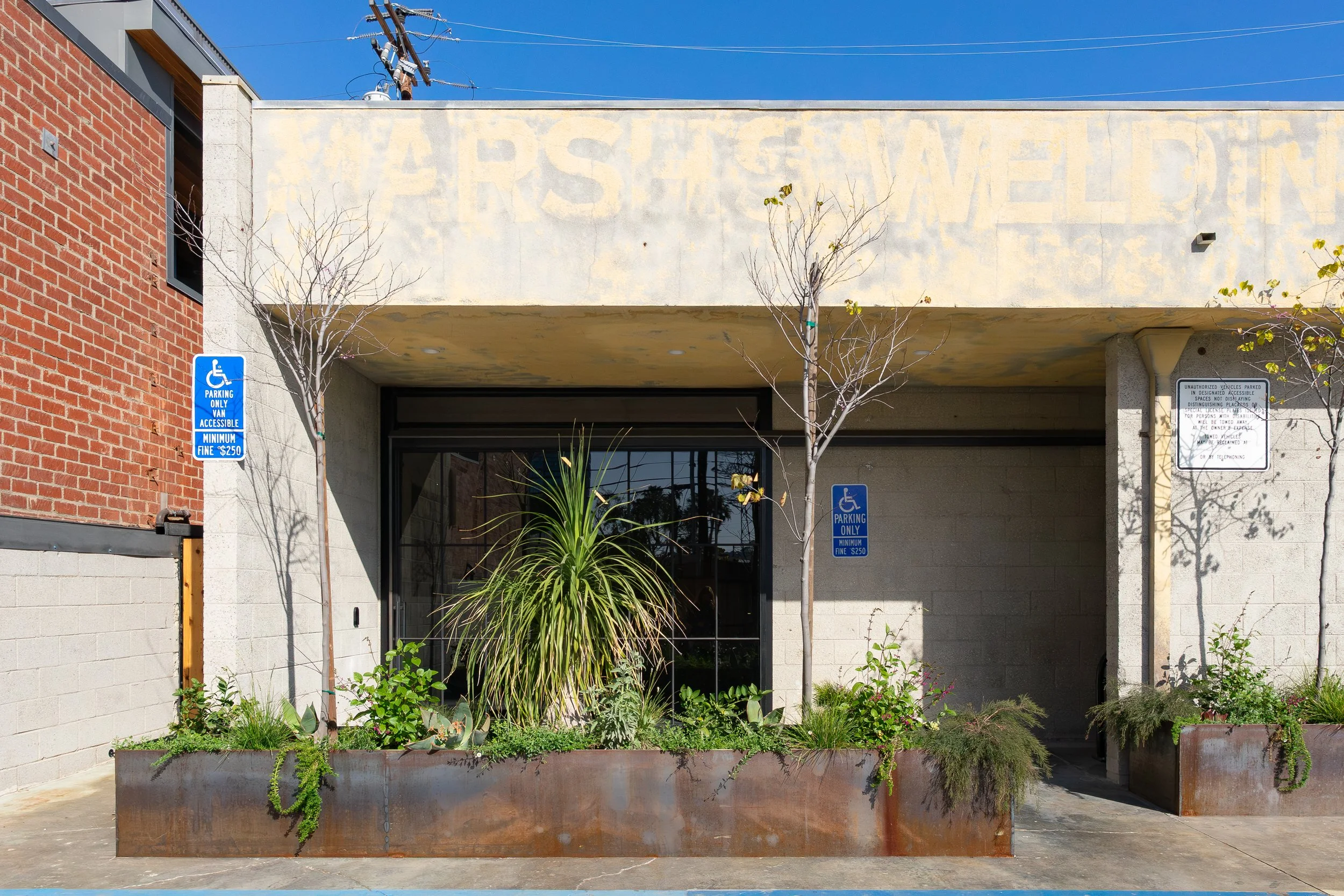 Street view of a building with a parking entrance, two trees in large metal planters, and blue handicapped parking signs.