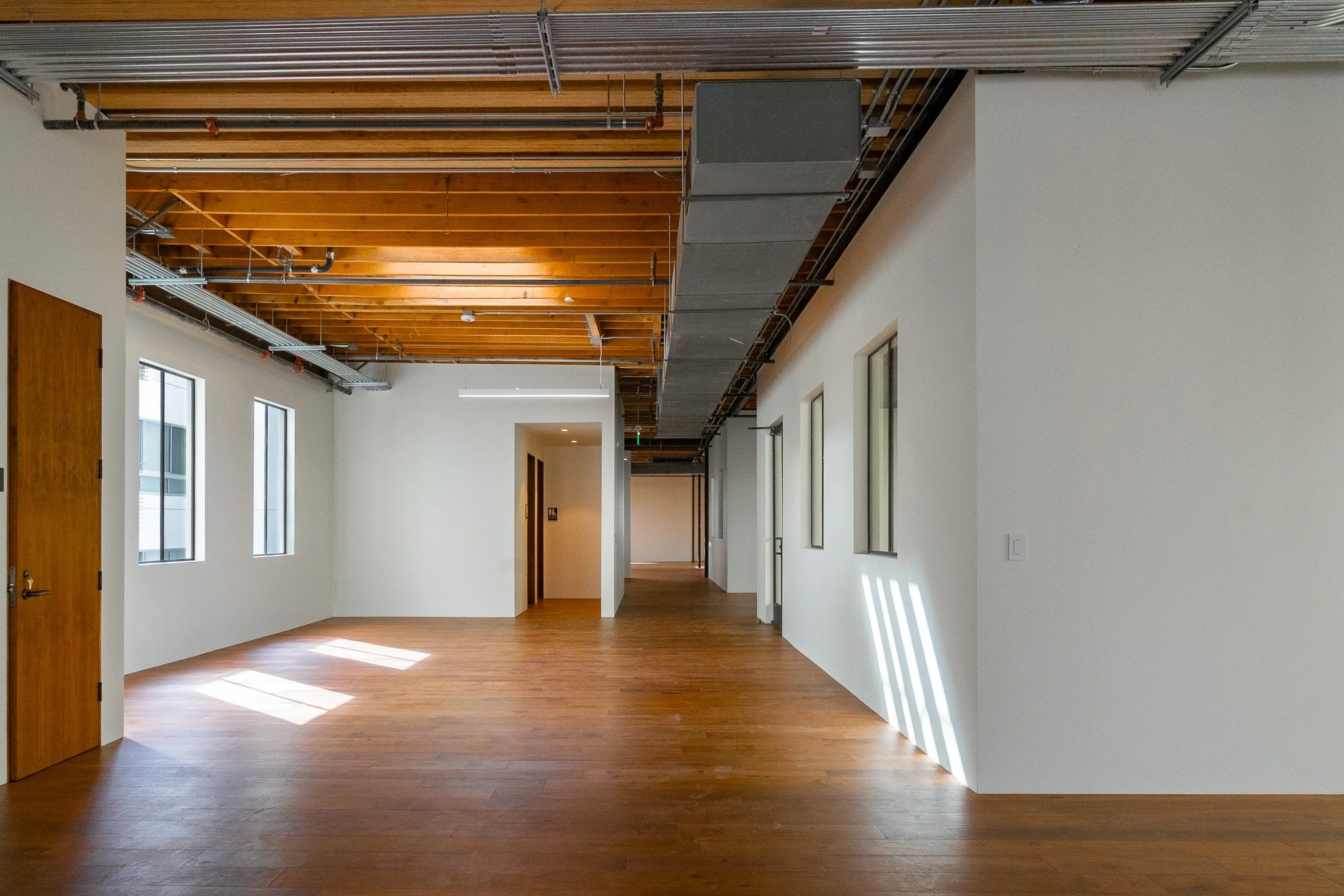 Empty modern interior space with wooden floor, white walls, windows, and exposed ceiling with wooden beams and metal ductwork.