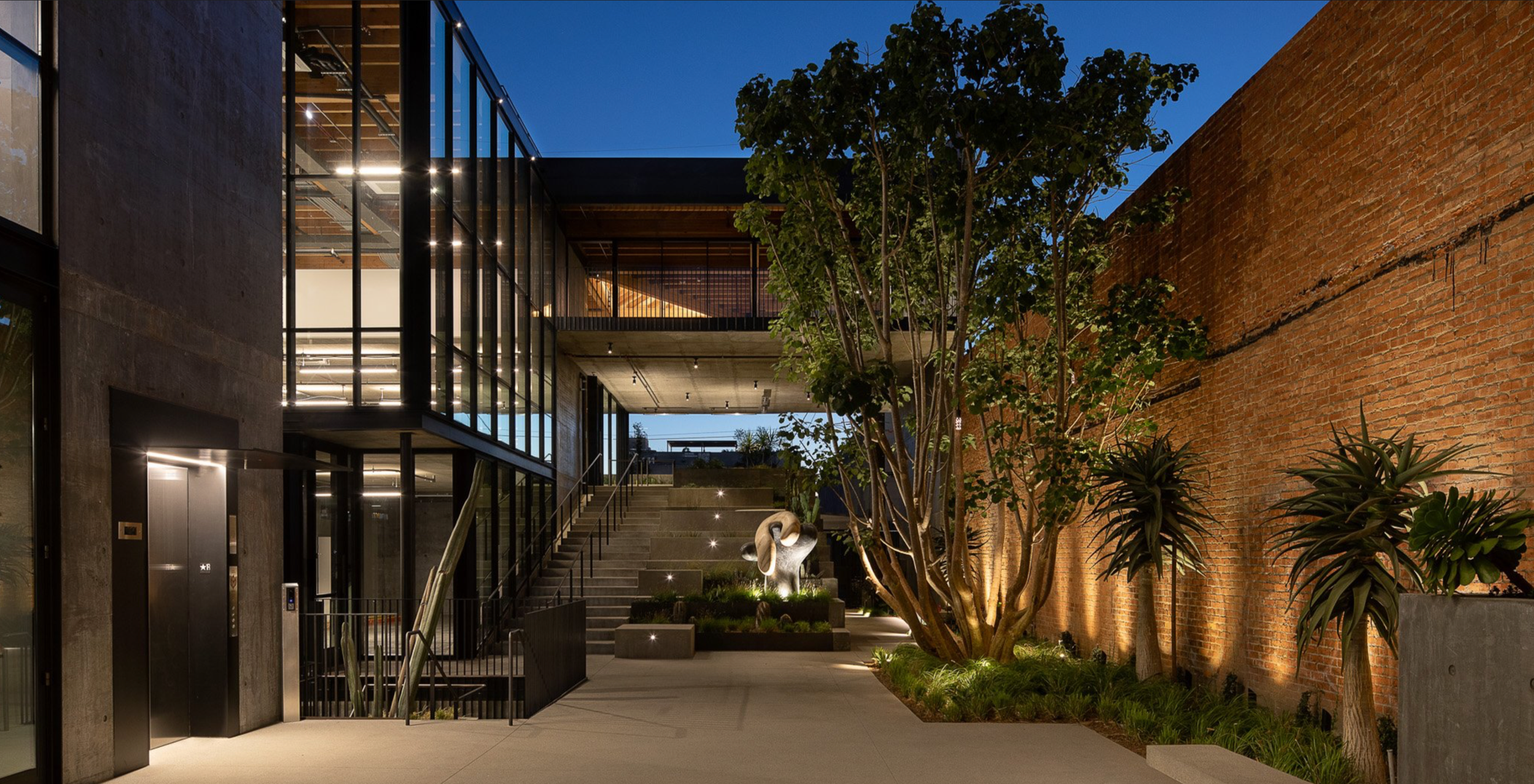 Modern urban building courtyard during evening with large glass windows, stairs, a sculpture of two intertwined figures, trees, and exterior lighting on a brick wall.