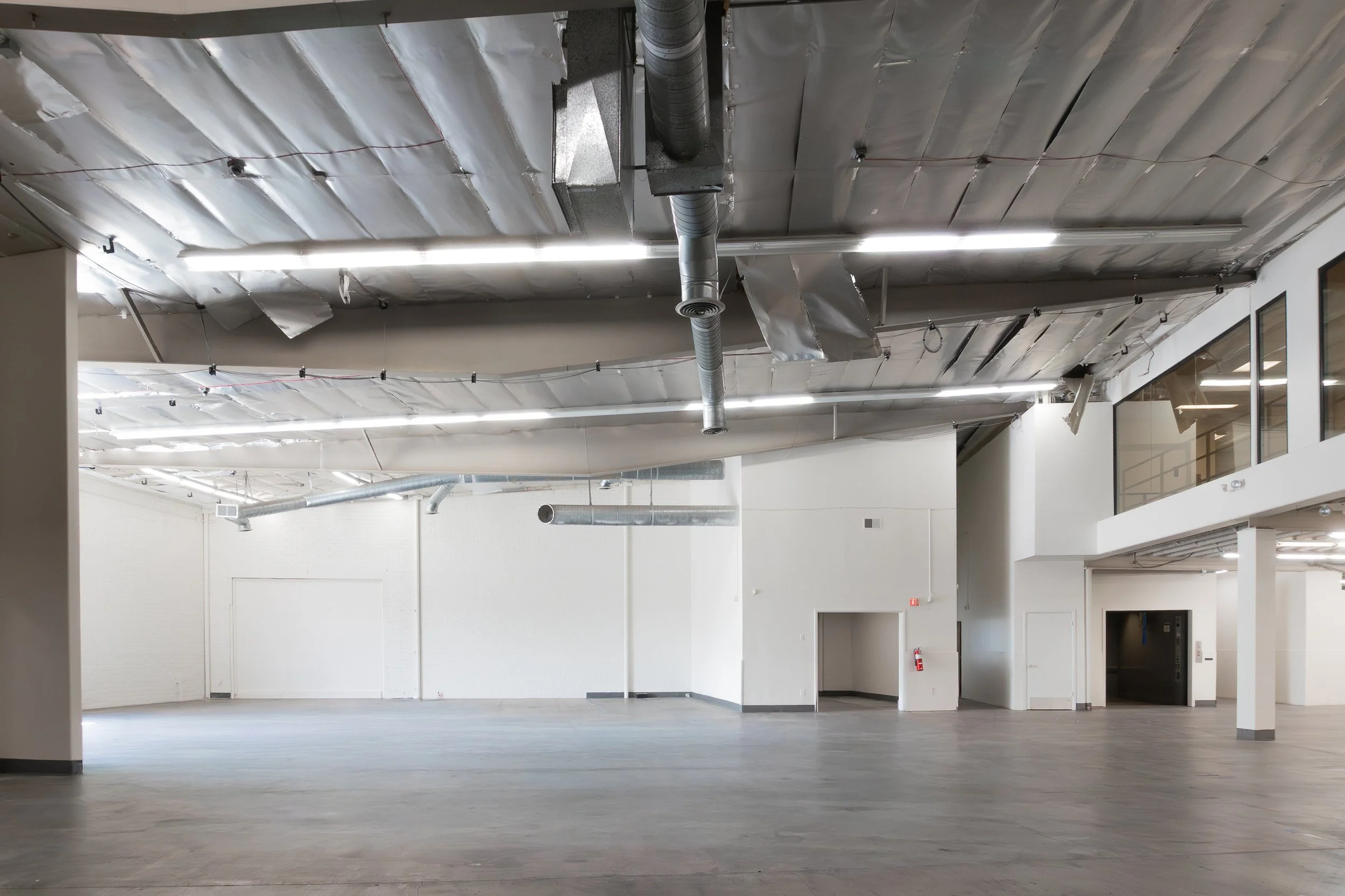 Empty industrial space with unfinished ceiling, exposed ductwork, concrete floor, white walls, and elevator door.