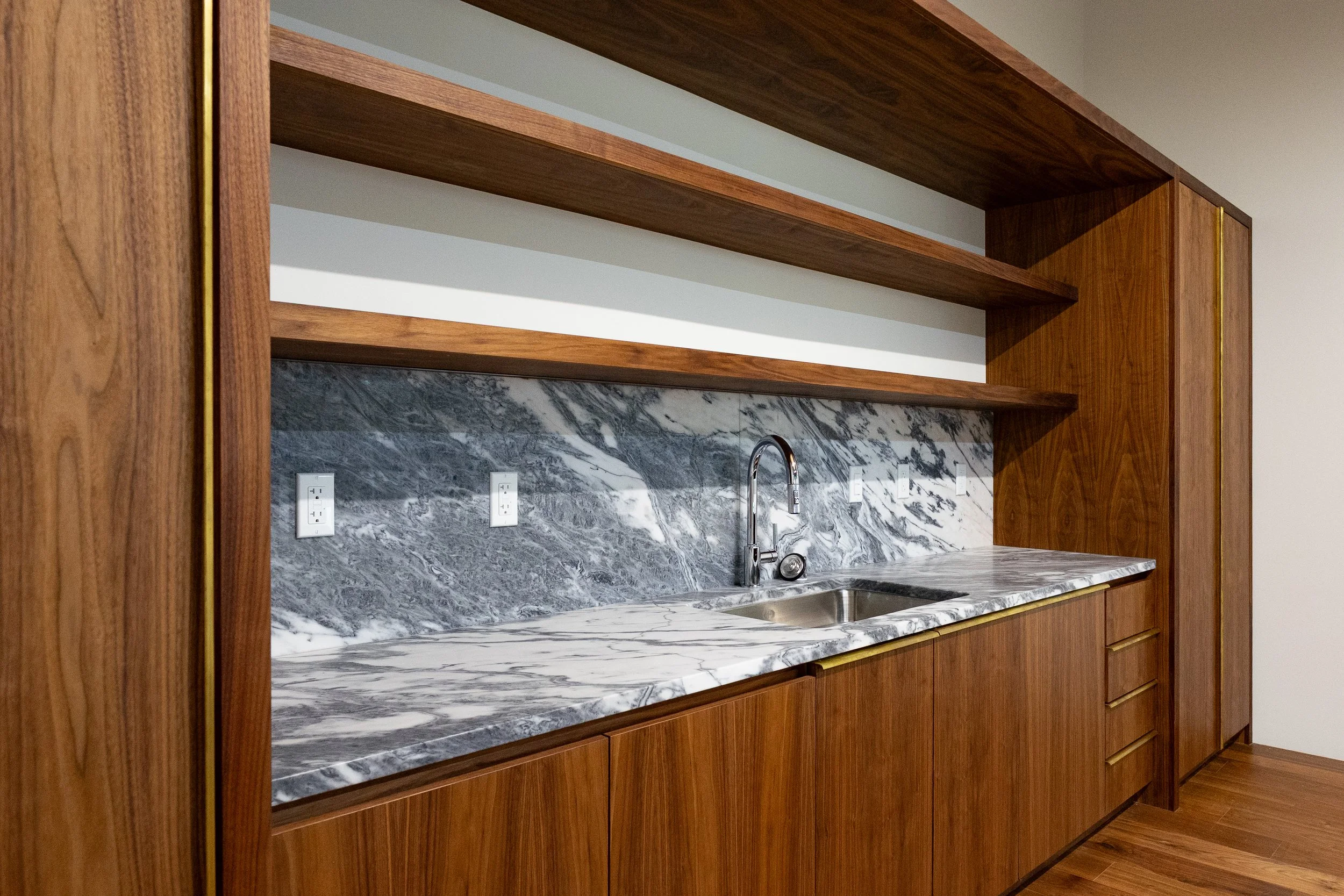 Kitchen with wooden cabinetry, marble countertop, marble backsplash, and a stainless steel sink with a faucet.