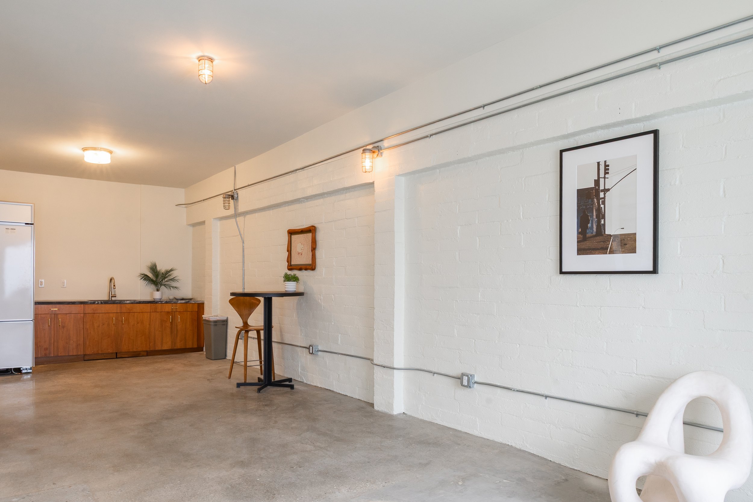 Minimalist interior space with white brick walls, concrete floor, wooden cabinetry with plants, framed artwork on the wall, and a white abstract chair.