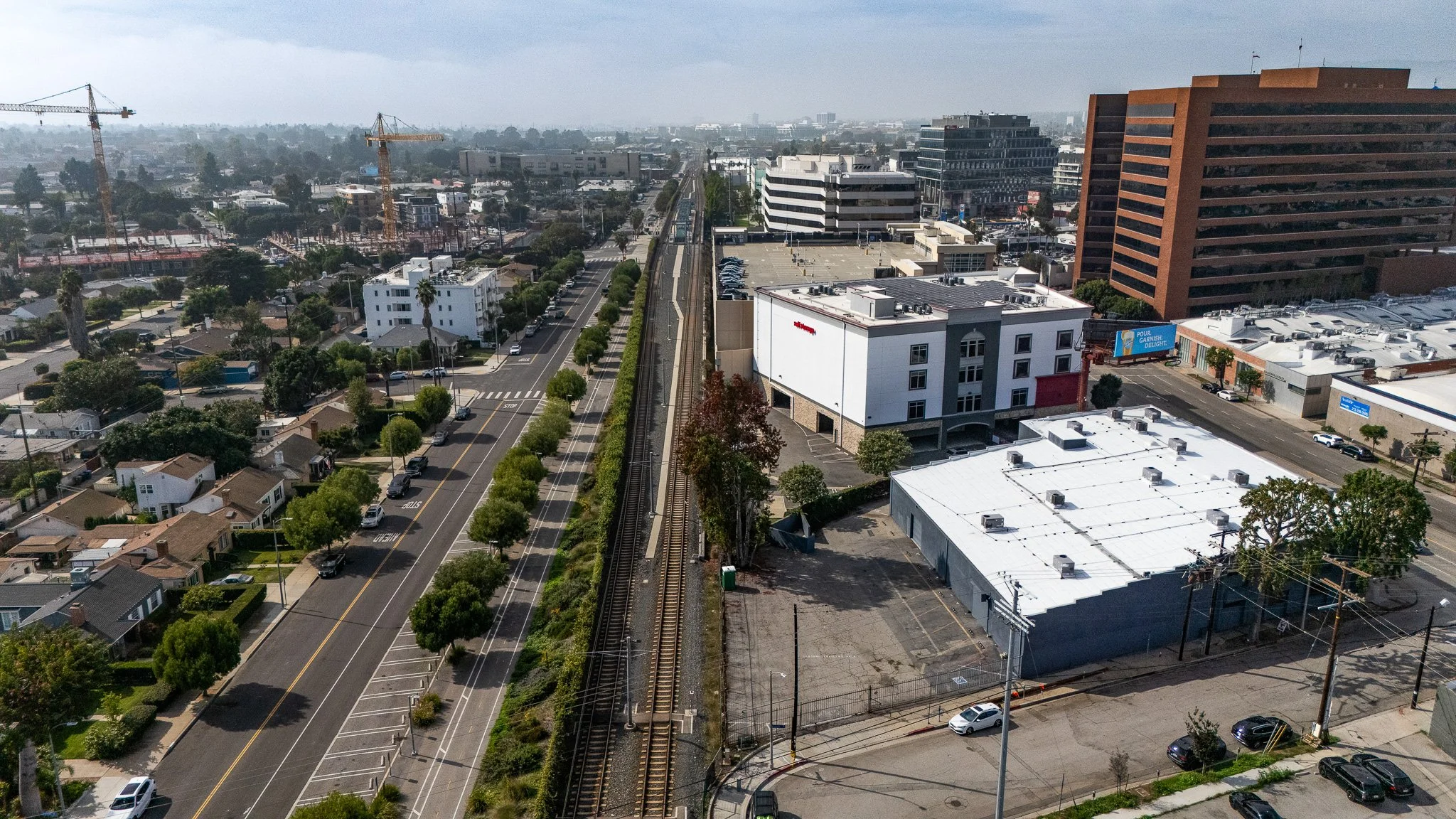 An aerial view of a city street with train tracks running parallel to a road lined with trees. Surrounding buildings include commercial and residential structures, with construction cranes visible in the distance and a large brown office building on the right.