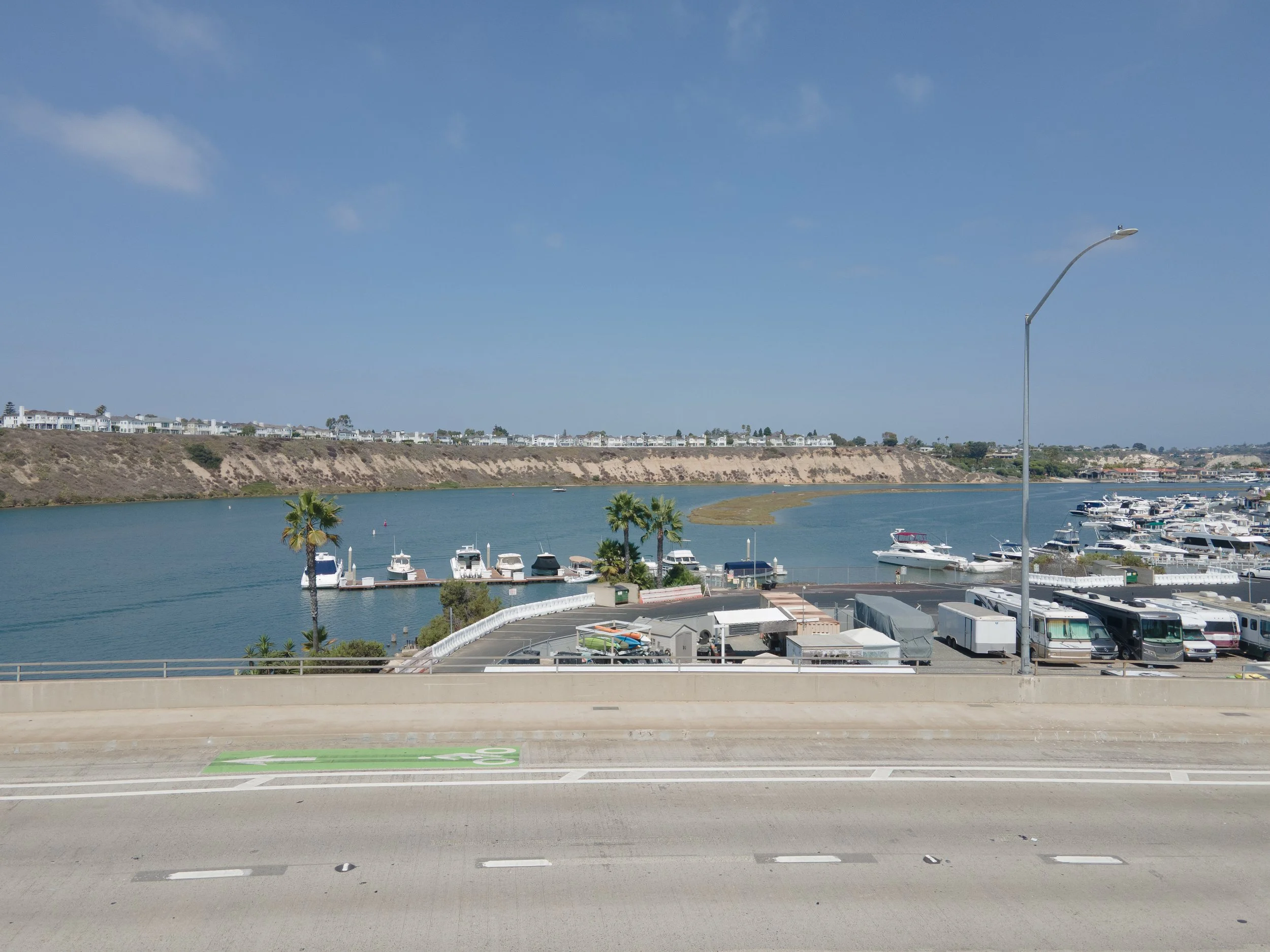 View of a marina with boats docked, a river, and a hillside with residential buildings in the background under a partly cloudy sky.