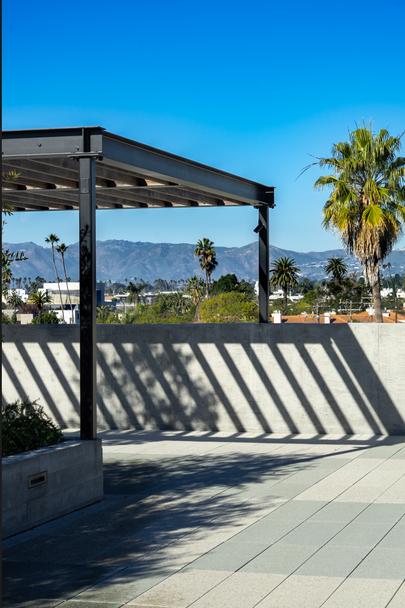 View of palm trees and mountains through a rooftop terrace with pergola and shadows on the ground, under a clear blue sky.