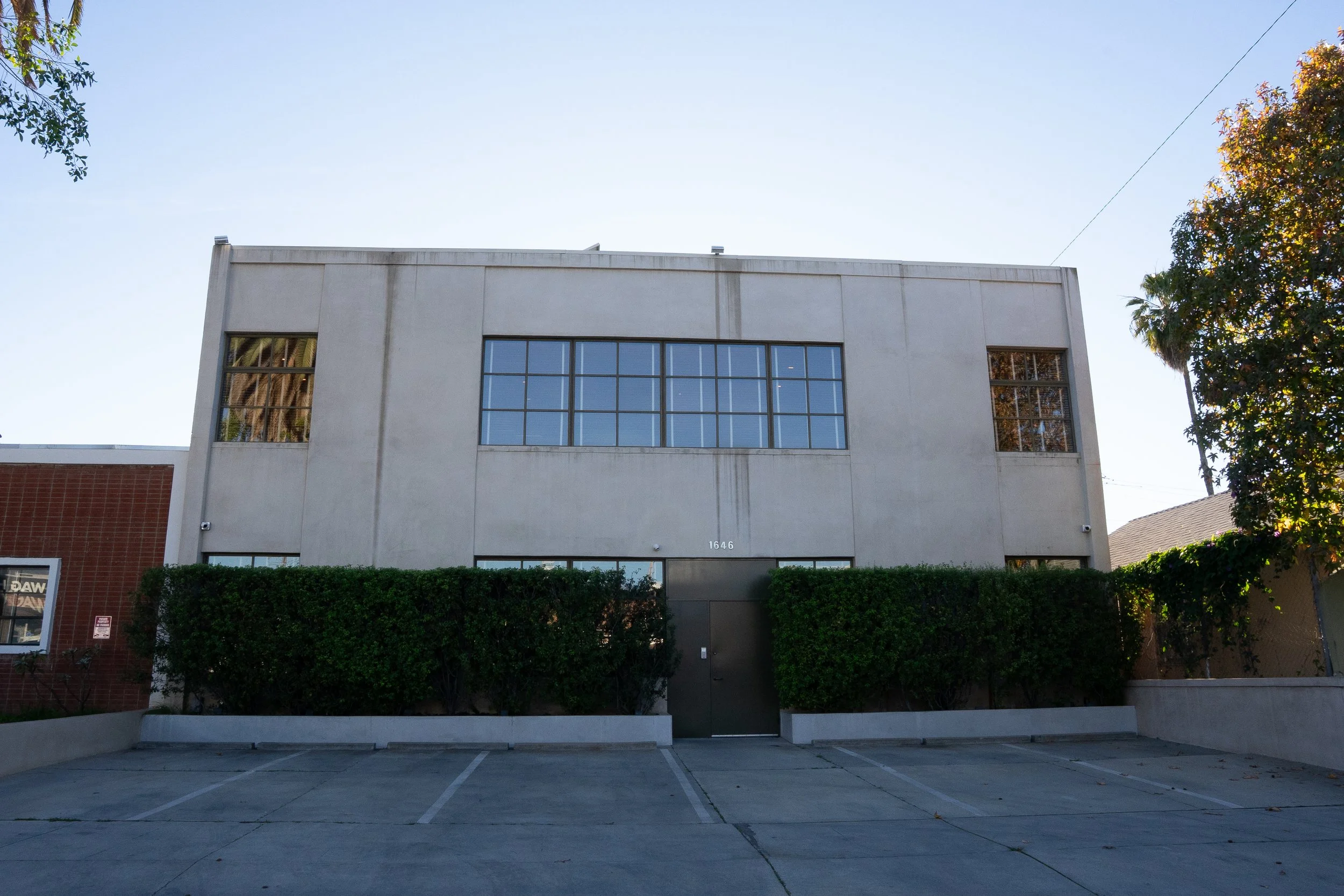 A two-story commercial building with a white facade, large rectangular windows on the upper floor, and a black door with the number 1646. There are neatly trimmed bushes in front and a parking lot in the foreground.