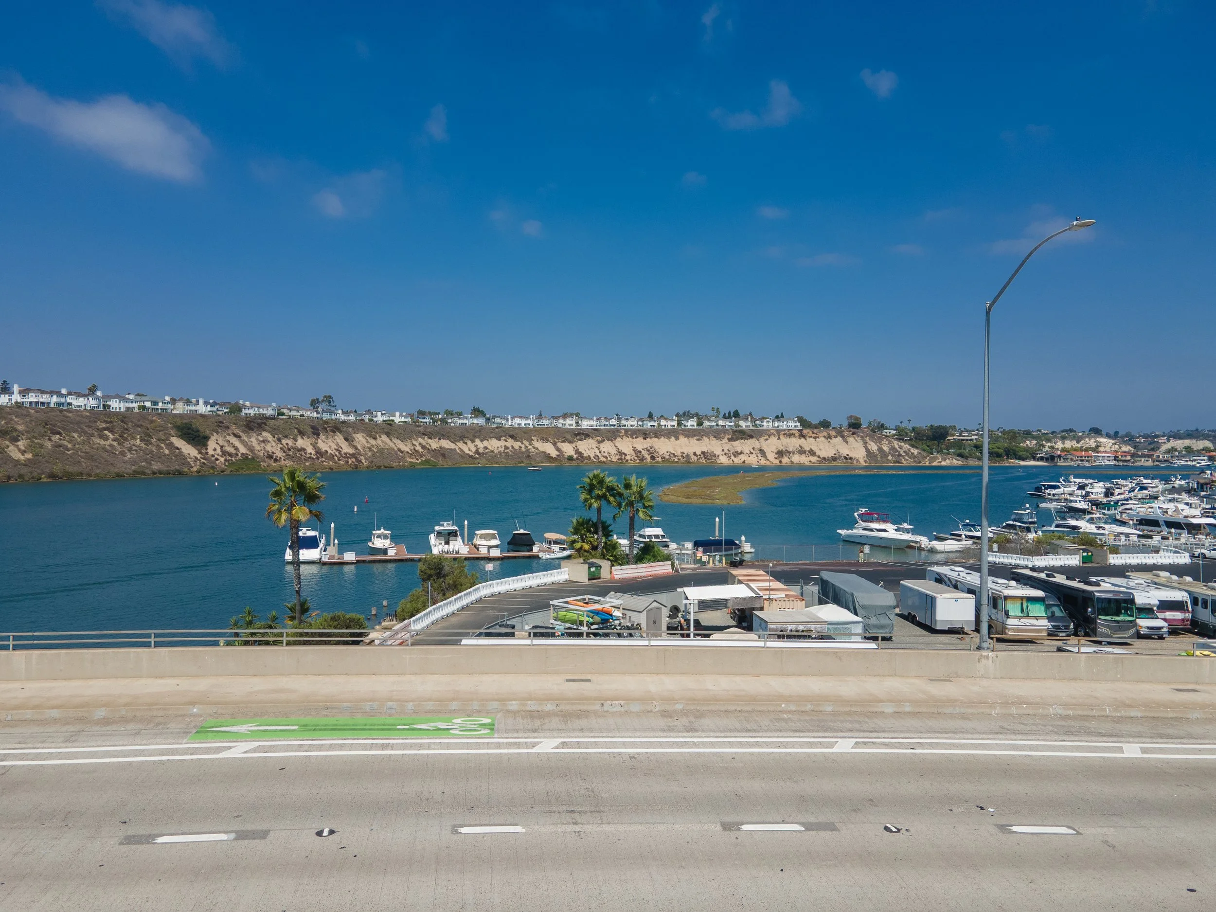 A marina with boats docked along the water, palm trees, a parking lot with RVs, and houses on a hill in the background under a partly cloudy blue sky.
