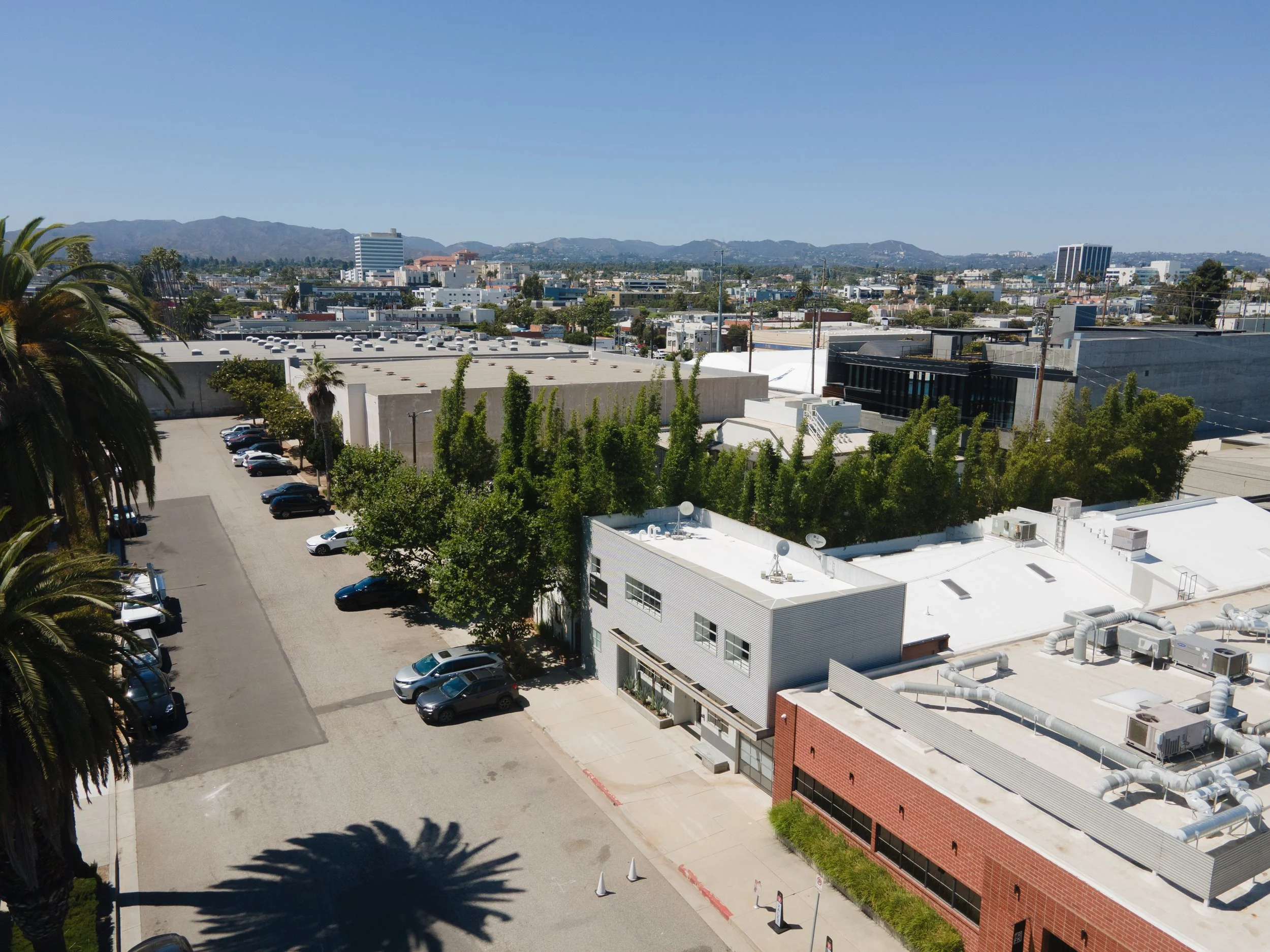 Urban cityscape with buildings, parking lots, trees, and mountains in the background under a clear blue sky.