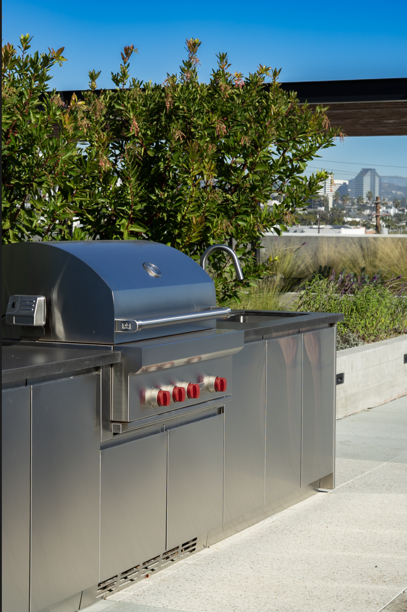 Stainless steel outdoor barbecue grill on a patio with a city skyline in the background under a clear blue sky.