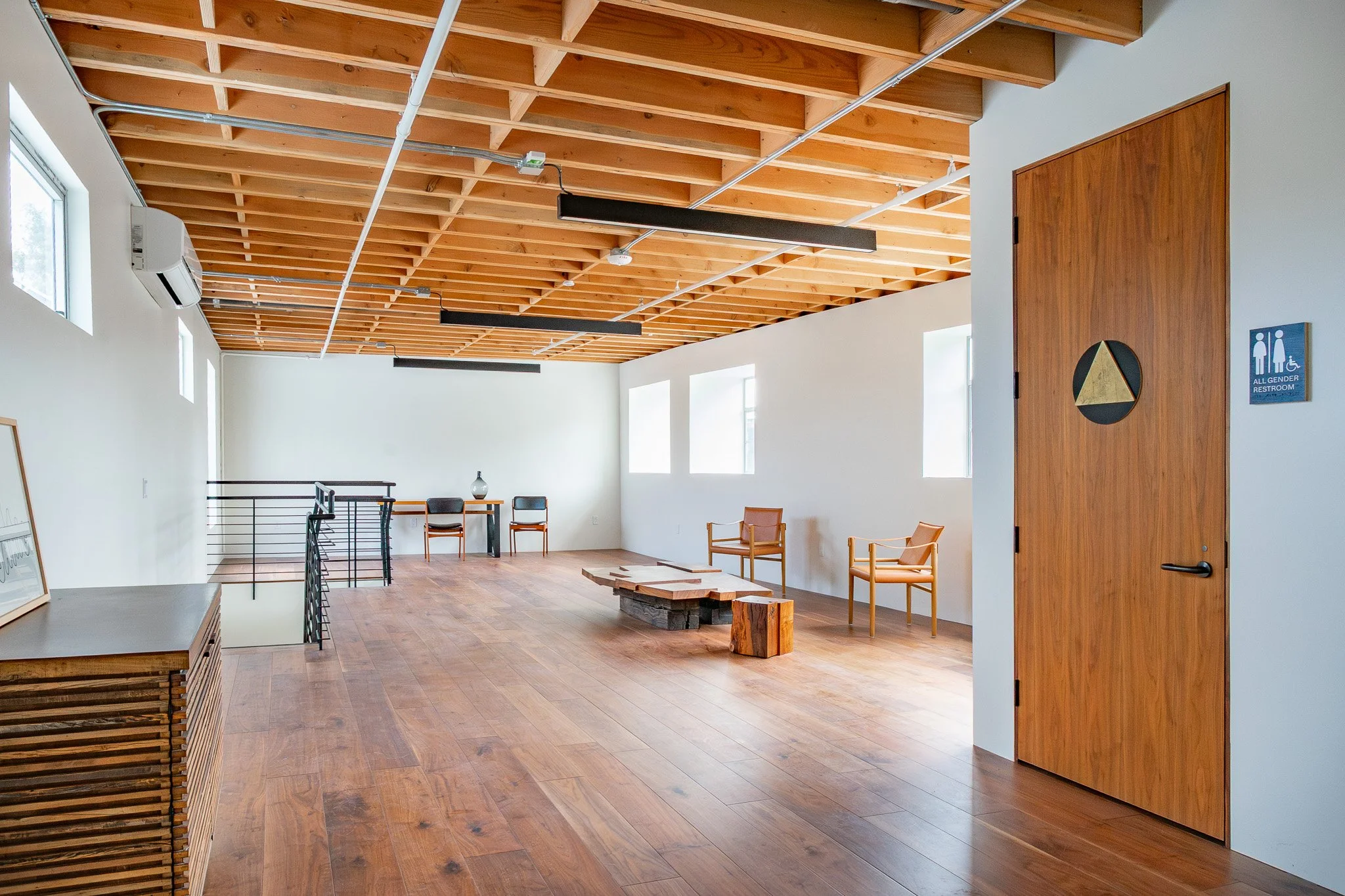 Interior of a modern, spacious room with a wooden ceiling, large windows, hardwood flooring, and minimalist furniture including chairs and a table. There is a restroom door on the right and some artwork on a side table.