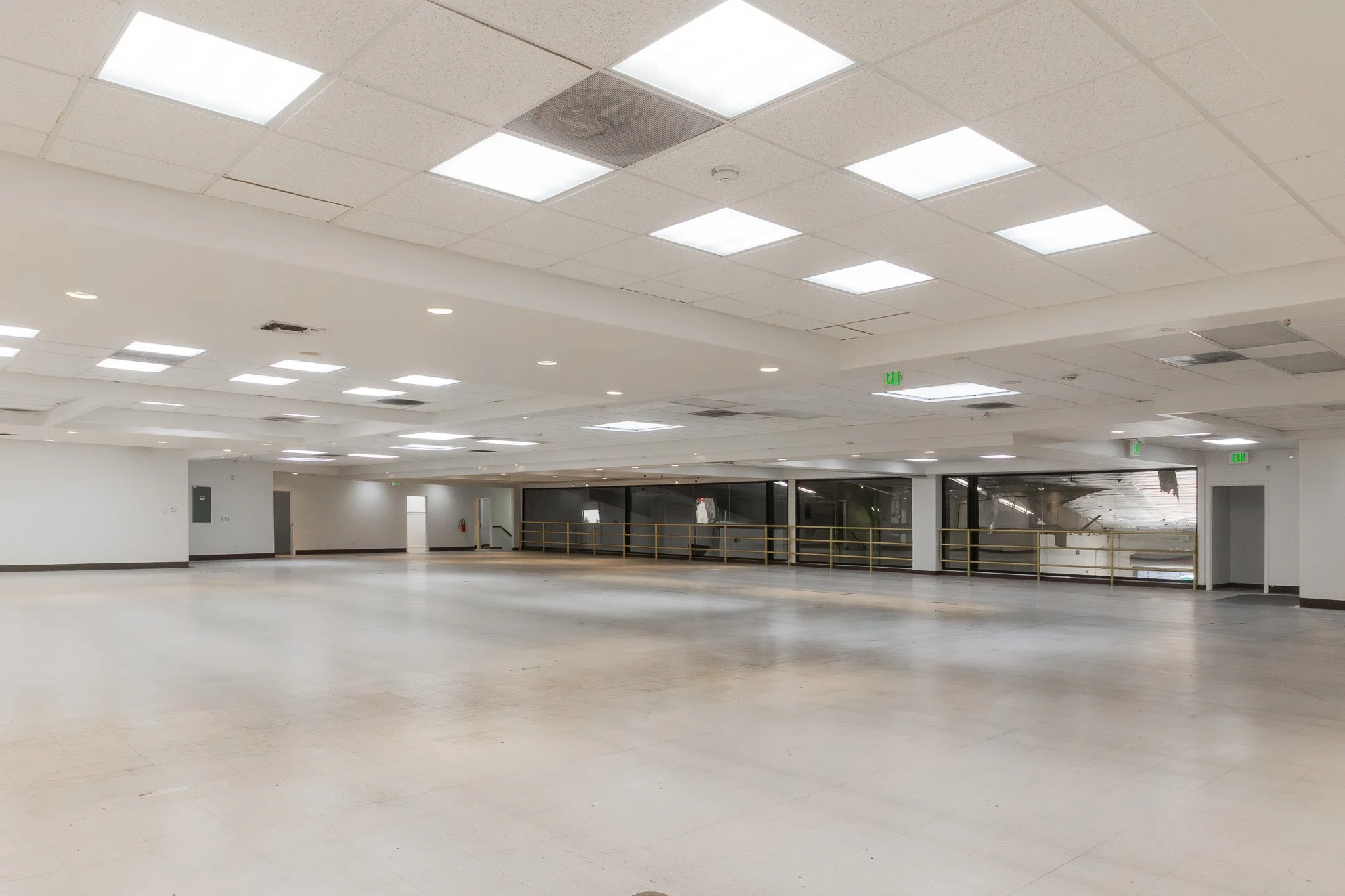 Empty indoor space with white walls, tiled ceiling with fluorescent lights, and large windows with black frames; the floor is bare and reflects light, with a railing along the windows and an emergency exit sign visible.
