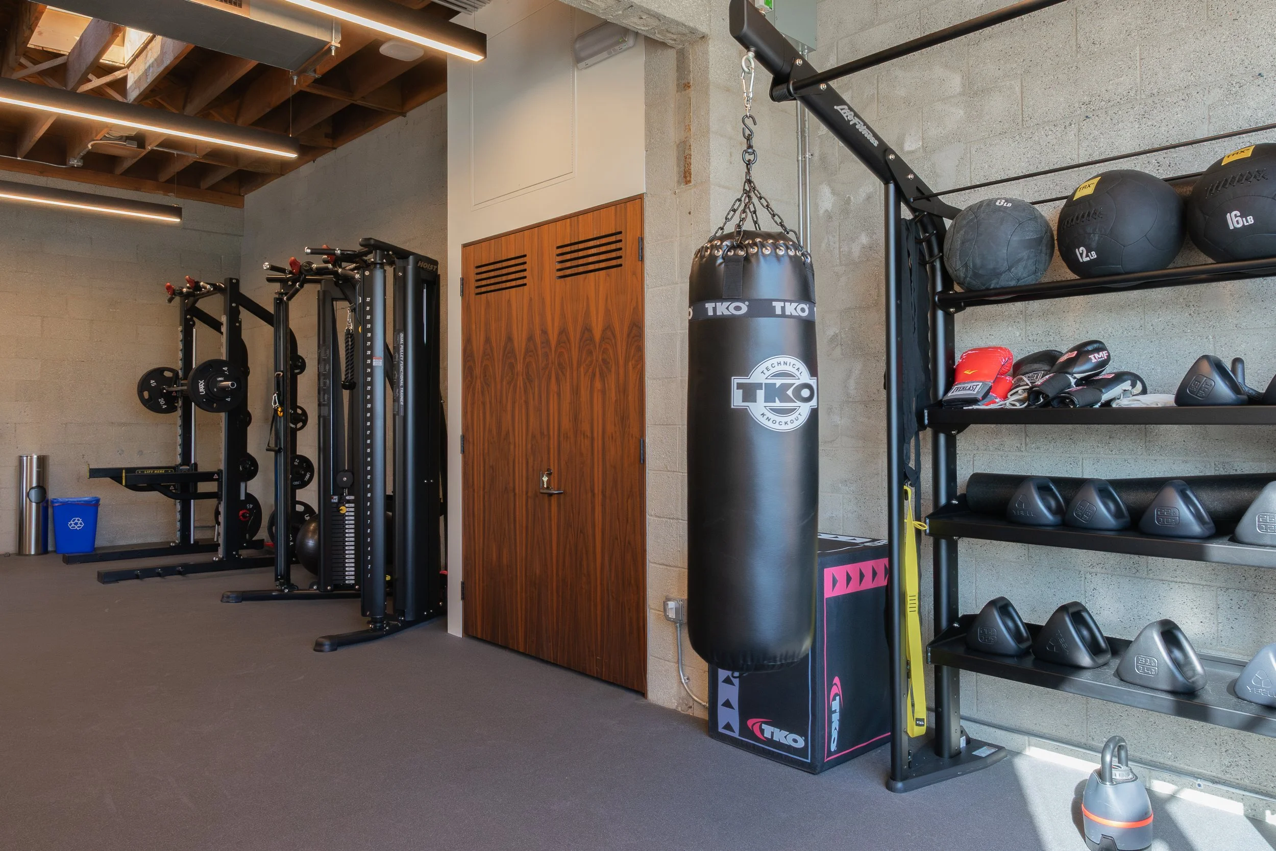 Gym interior with workout equipment including a punching bag, weight racks, medicine balls, boxing gloves, and kettlebells.