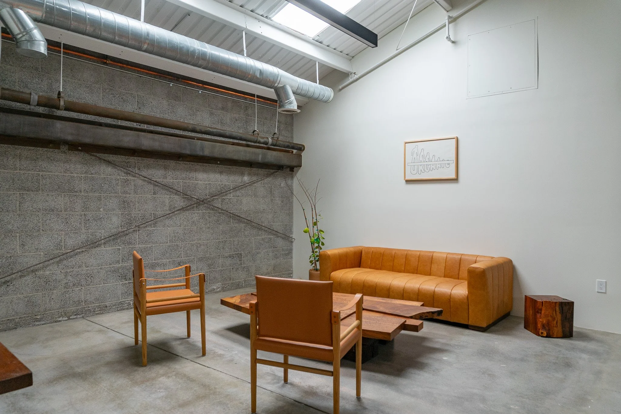 Sparse modern lounge with tan leather sofa, two wooden chairs with tan cushions, a rectangle wooden coffee table, potted plant, minimalist artwork on white wall, and gray concrete floor. Exposed ductwork on ceiling.