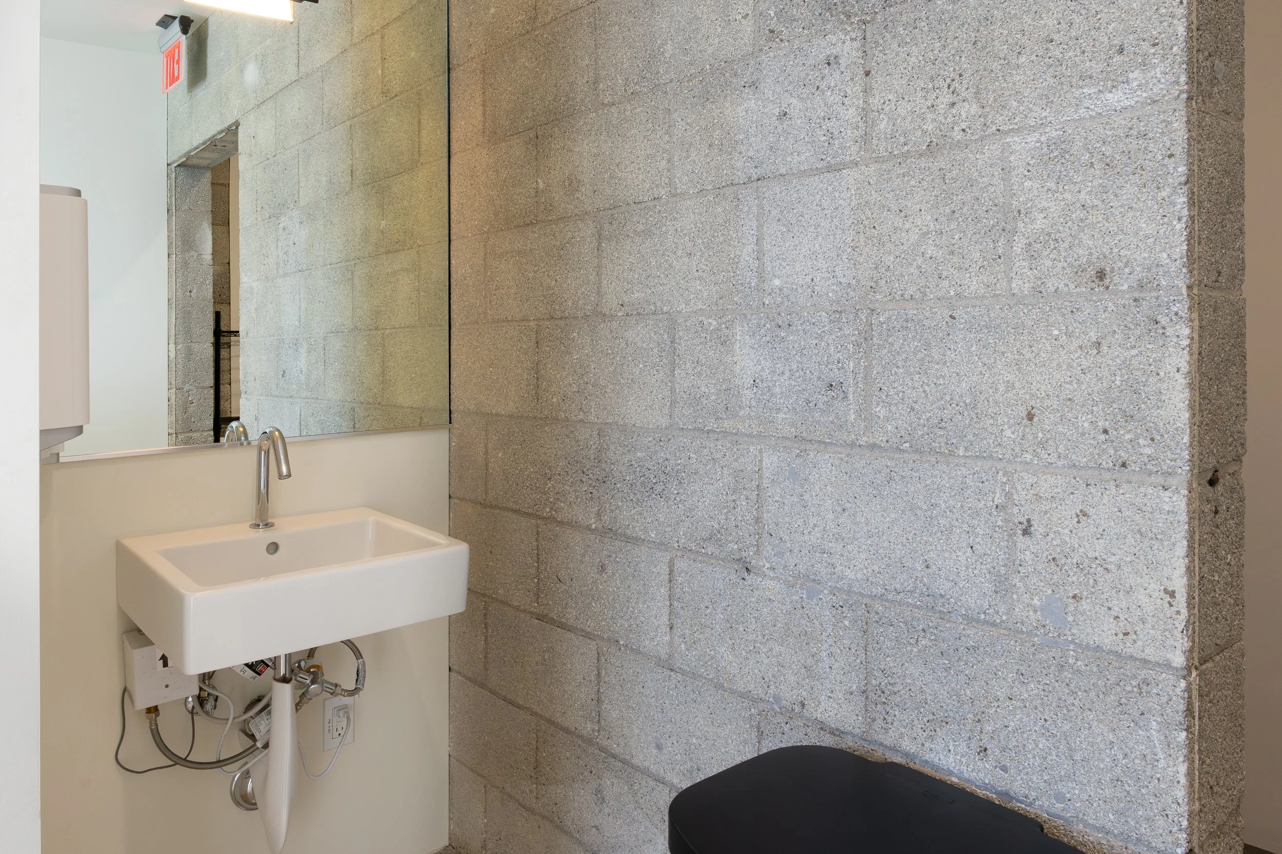A small utility sink with exposed plumbing and a faucet, set against a beige painted wall and a large mirror. To the right, a concrete cinder block wall is visible. There is a black bench or seat at the bottom right of the image.