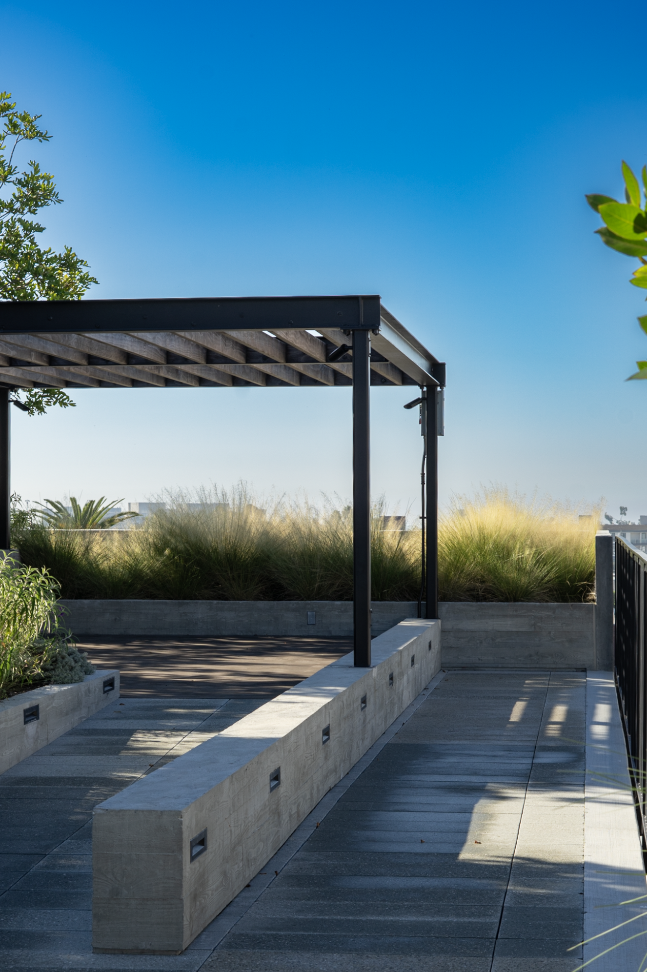 Rooftop terrace with a modern seating area, concrete bench, wooden pergola frame, lush grass landscaping, and clear blue sky.