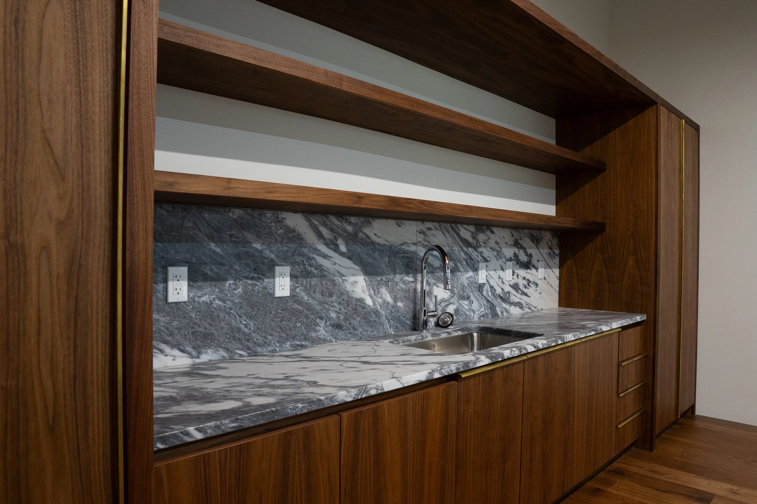 A kitchen with a marble countertop and backsplash, wooden cabinets, a stainless steel sink, and electrical outlets.