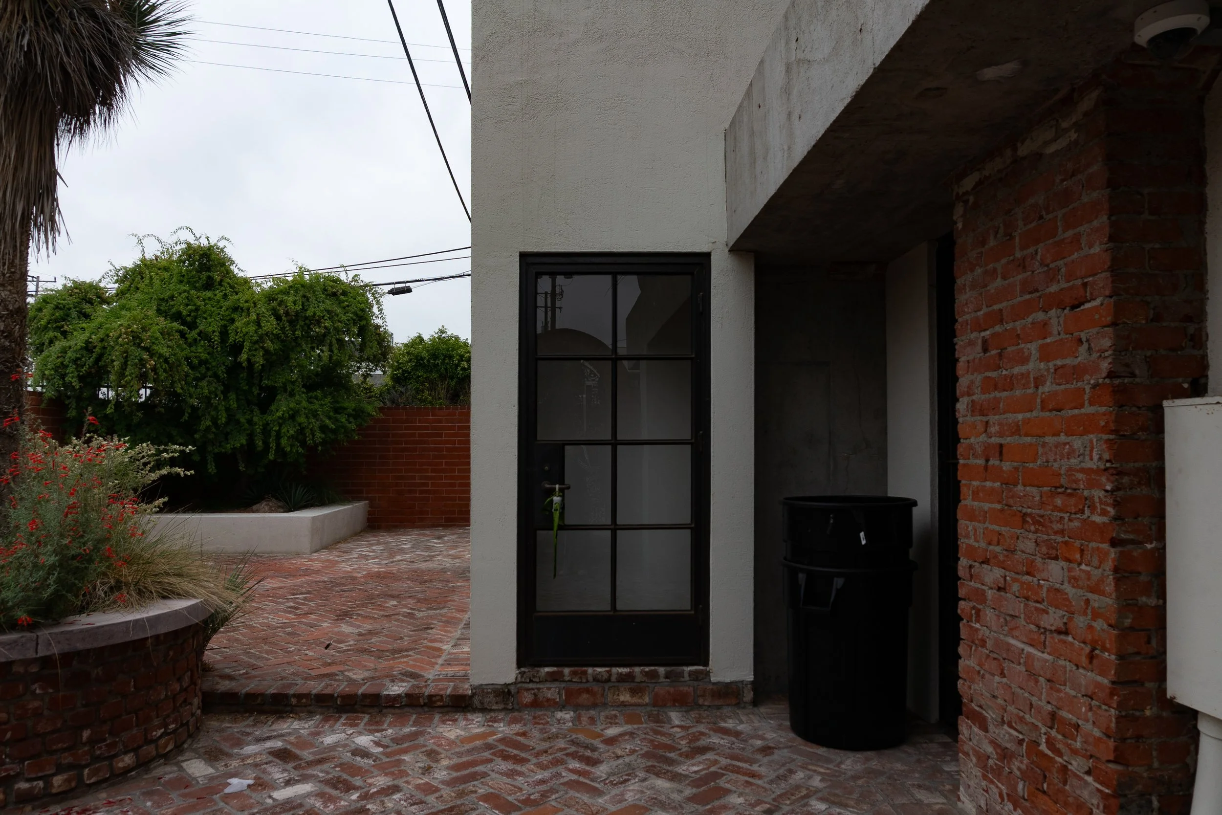Photo of a small outdoor patio area with red brick paving, an area with plants, and a building corner with a black-framed glass door. The building has a white exterior wall and exposed brickwork. A black trash bin is under a covered section next to t