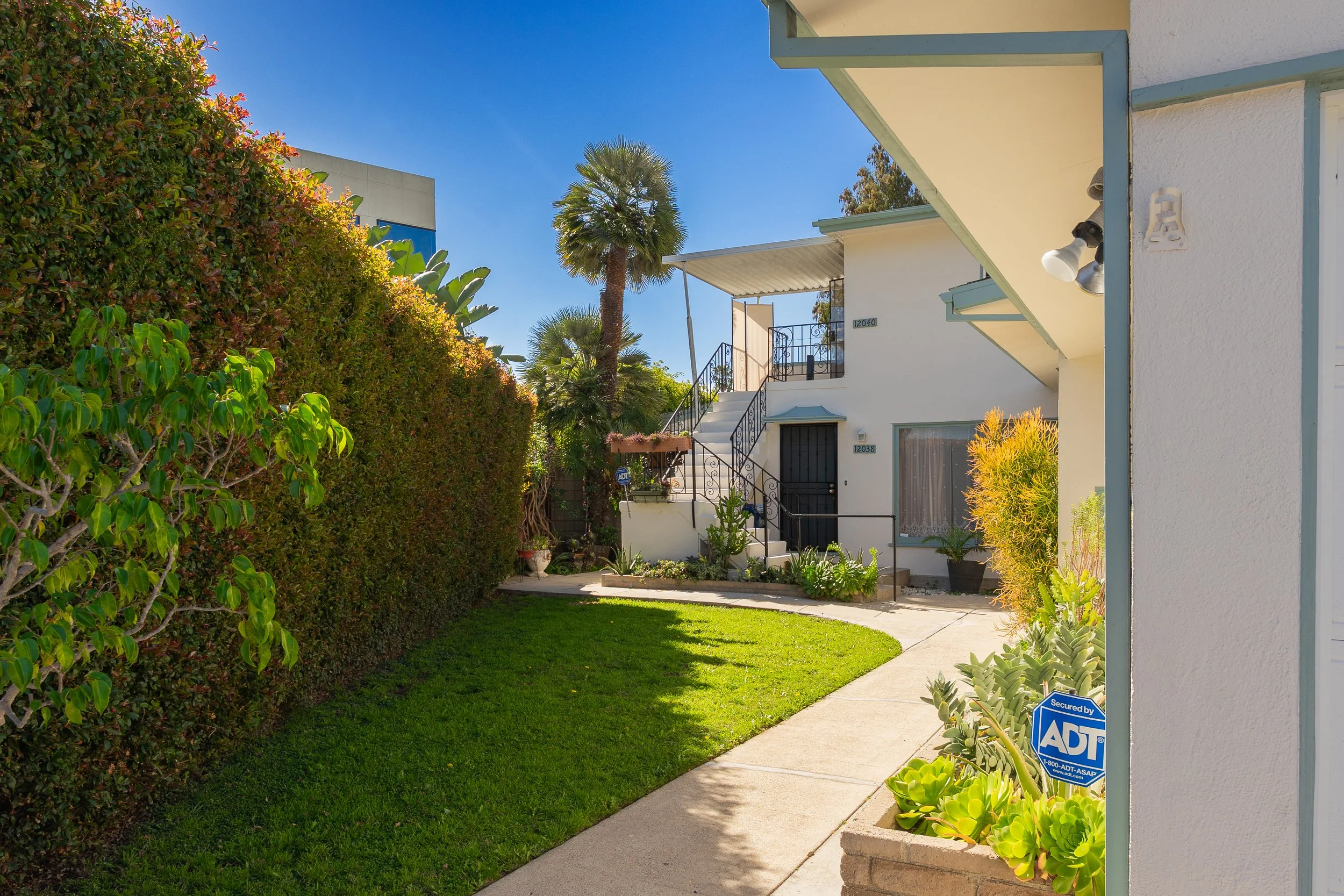 View of a sunny residential front yard with a green lawn, a concrete walkway, a tall hedge on the left, potted plants, and a white two-story house with stairs leading to the second floor in the background. A palm tree is visible behind the house, and