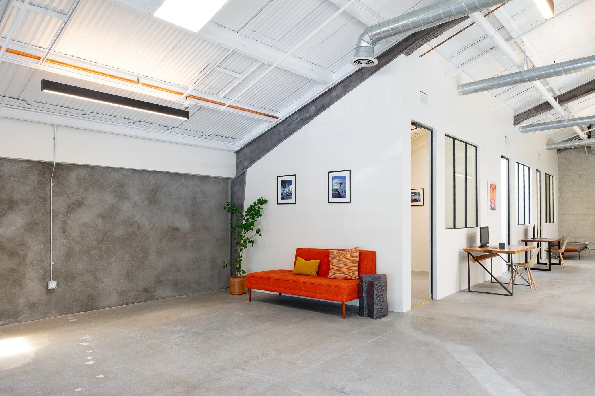 Modern interior with a concrete wall, an orange sofa with yellow and orange pillows, a green plant in a large pot, framed artwork on the white wall, next to a workspace with a desk and chair, and large windows letting in natural light.