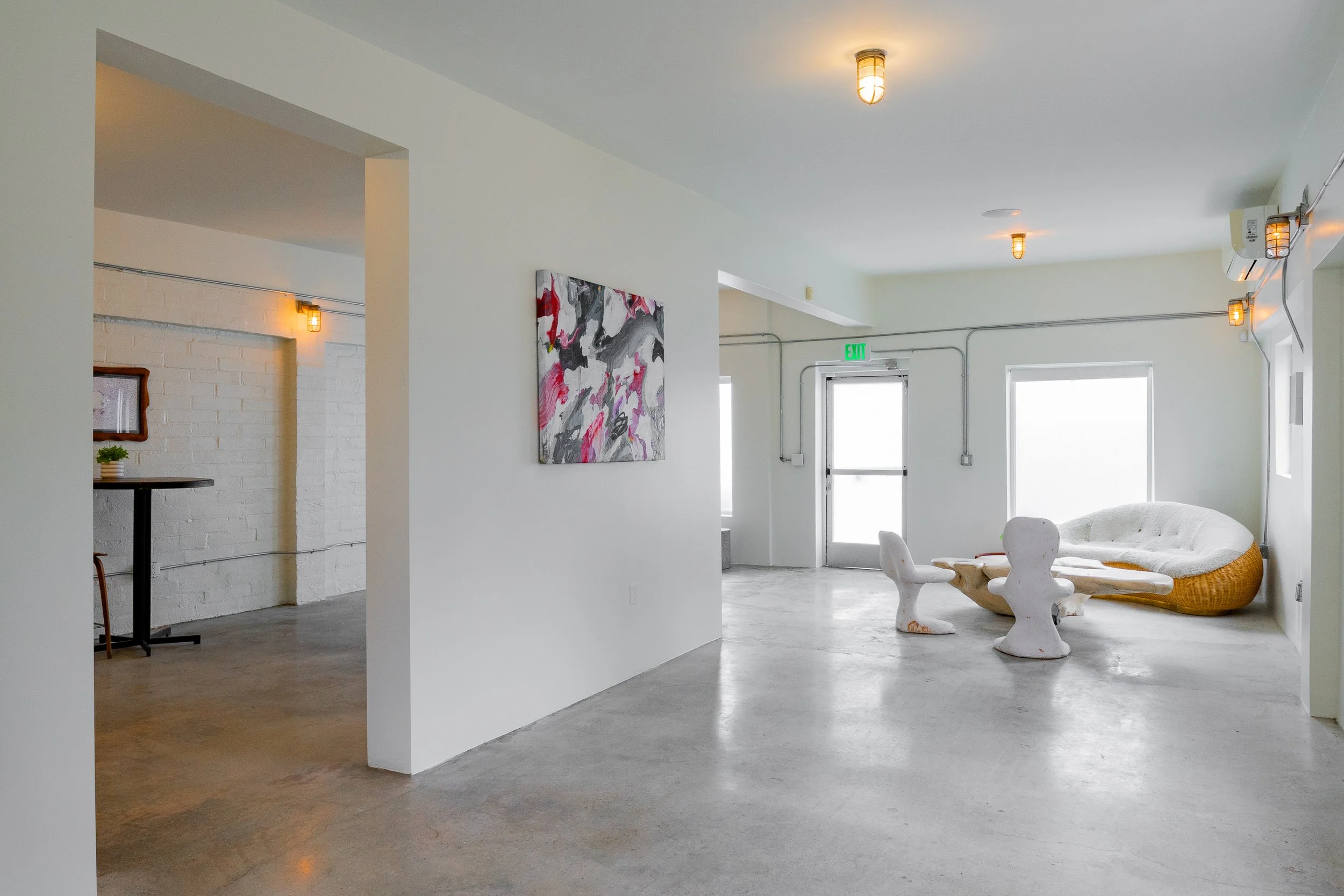 Minimalist interior with white walls and concrete floor, art on wall, seating area with white abstract chairs and a curved white lounge chair near windows.
